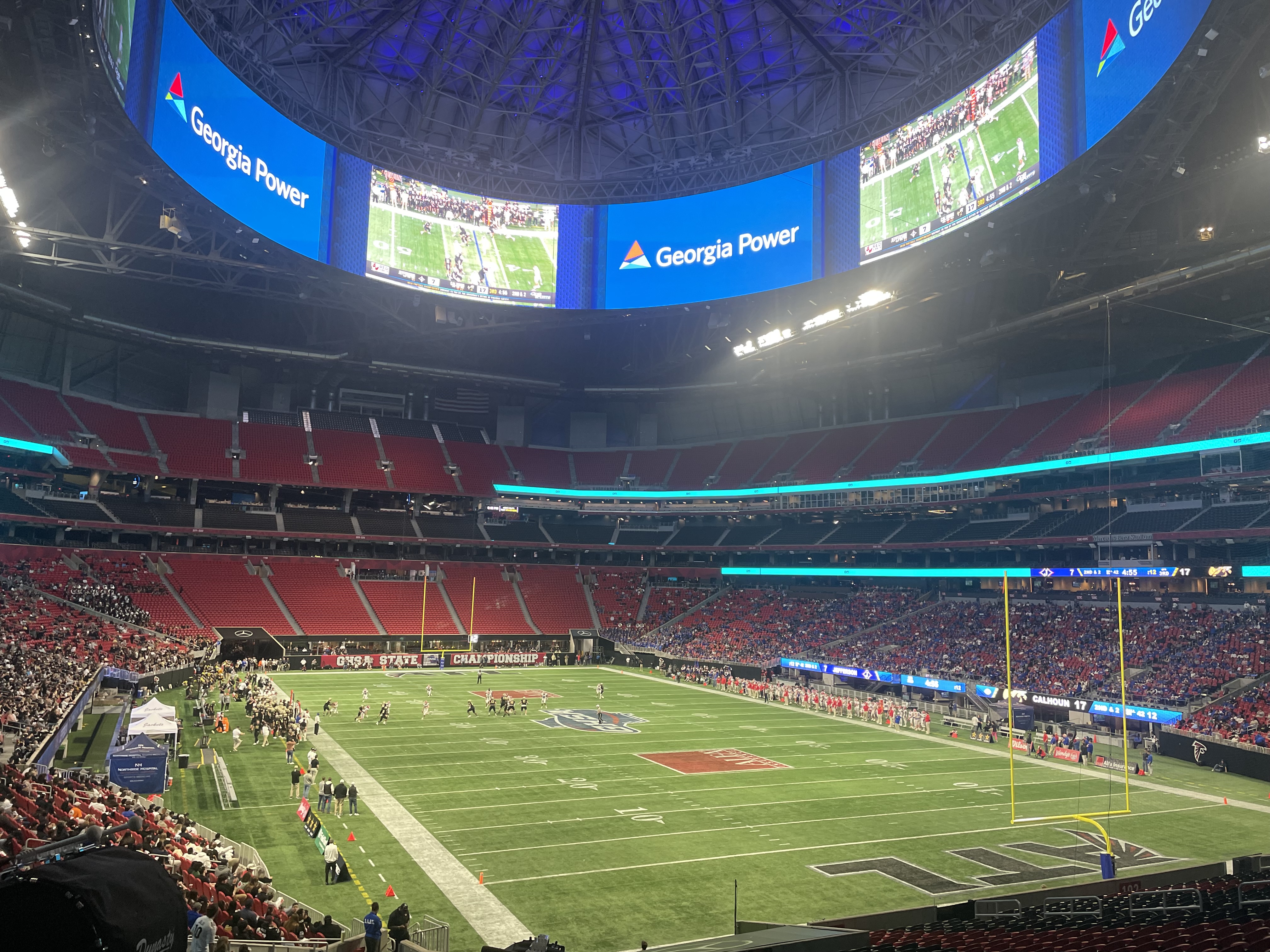 Football game in progress inside a large domed stadium with red seating and video boards.