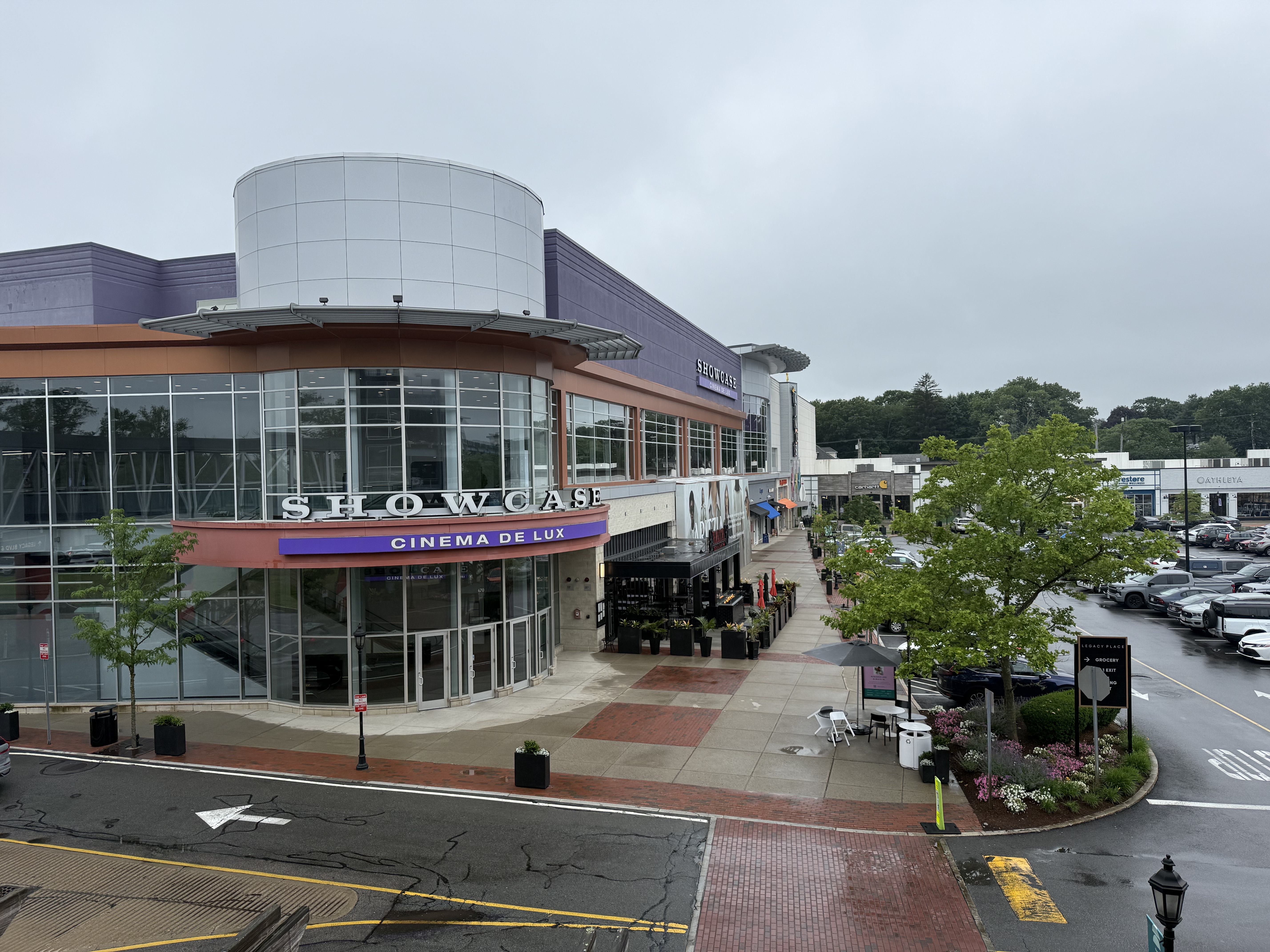 Street-level view of a cinema entrance at an open-air shopping center with a parking lot and retail stores.