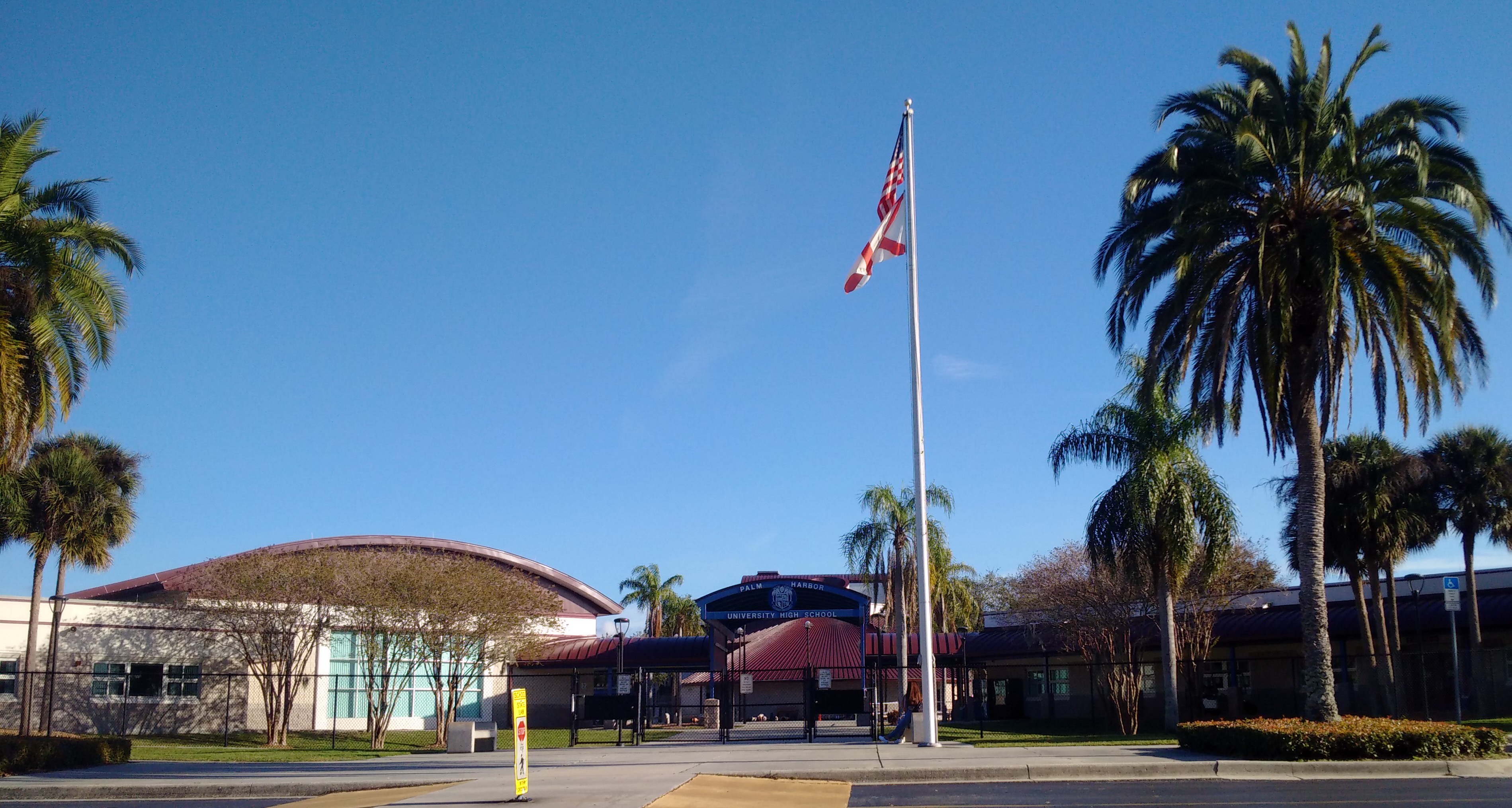 Exterior of a high school campus with palm trees, a flagpole, and a curved-roof gymnasium under a blue sky.