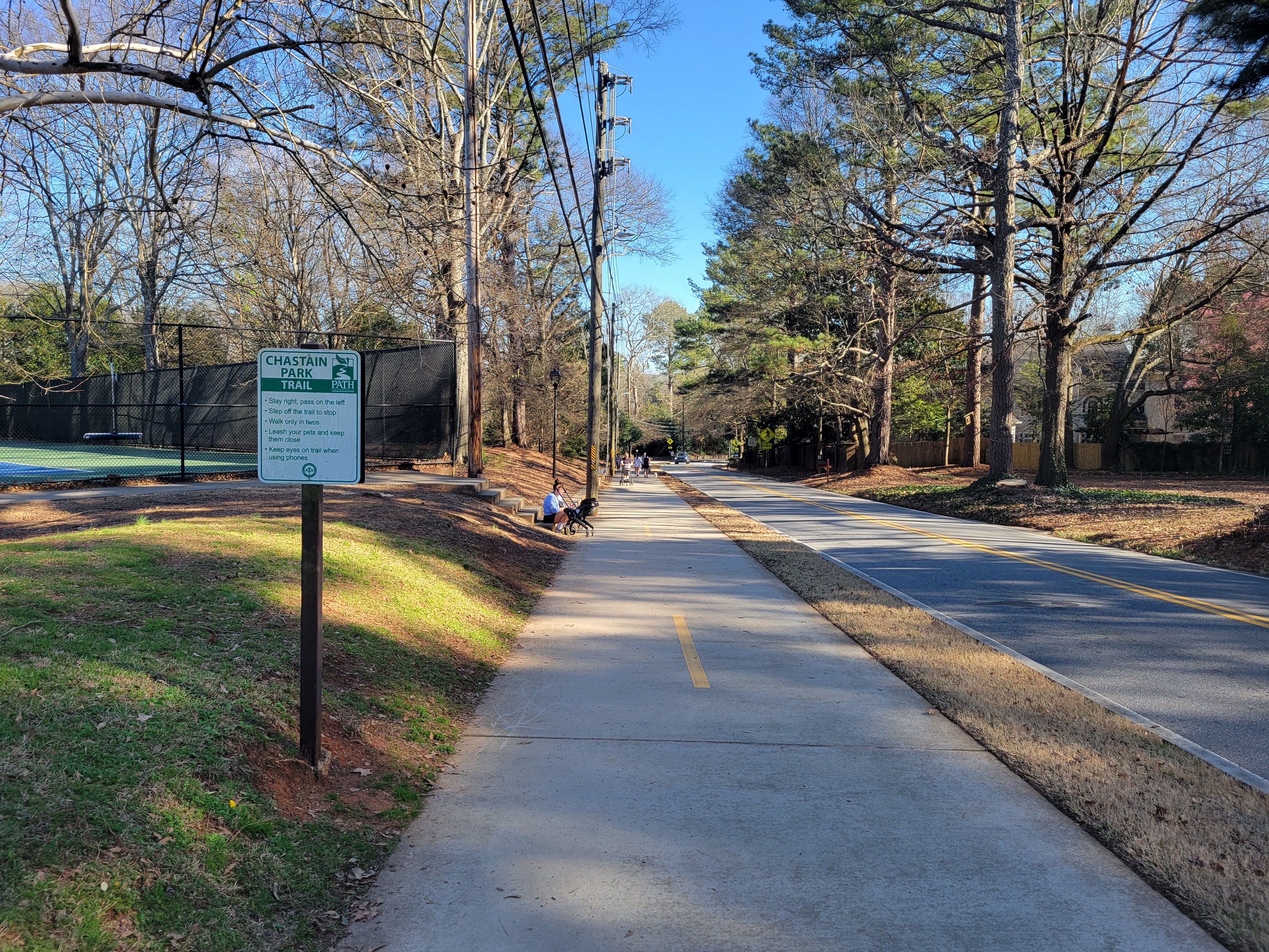 Paved sidewalk alongside a tree-lined road at Chastain Park, with a green park sign visible.