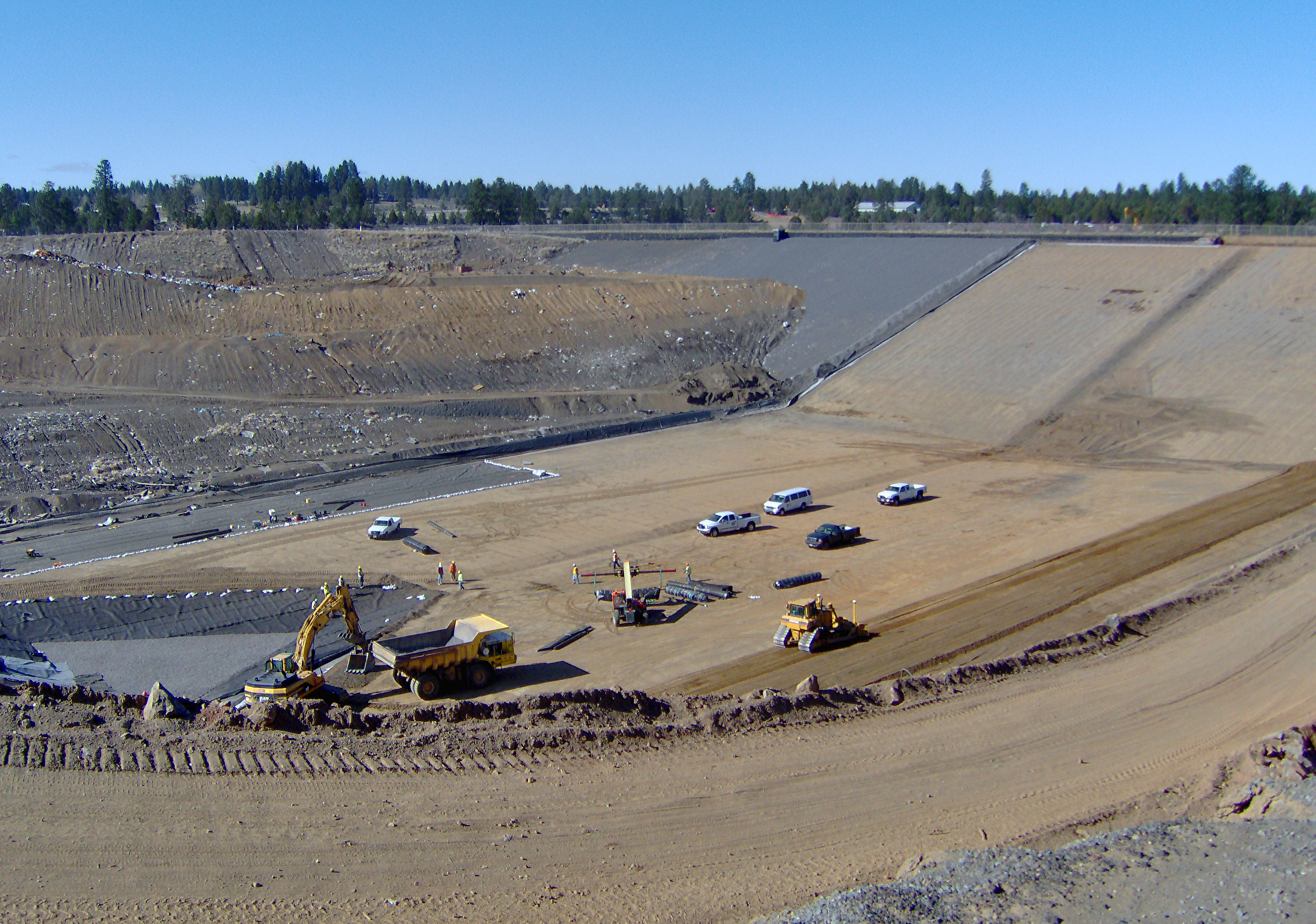Heavy equipment and dump trucks on a large graded earthen pad at a landfill site.