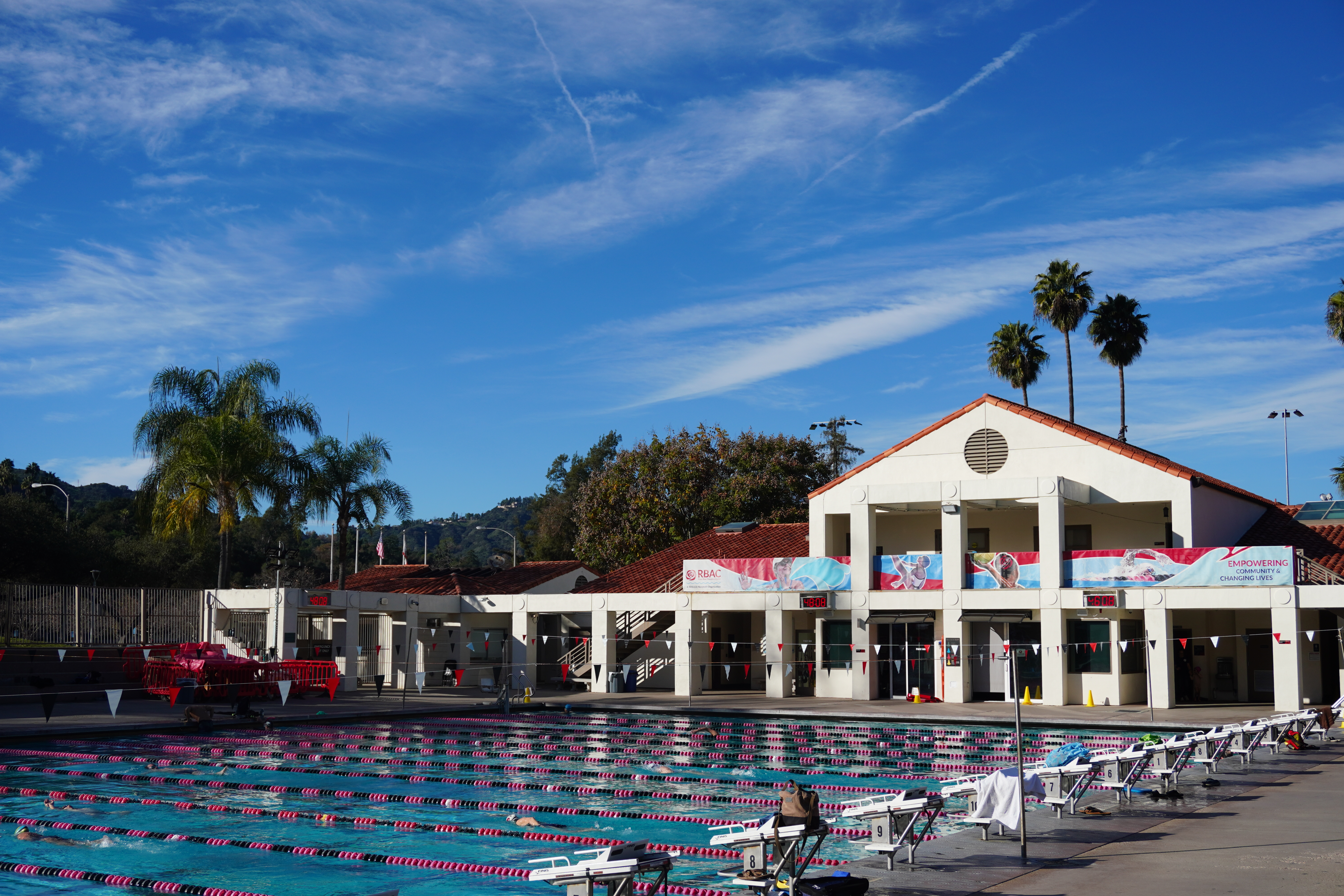 Outdoor aquatics center with competition pool lanes, white building, and palm trees.