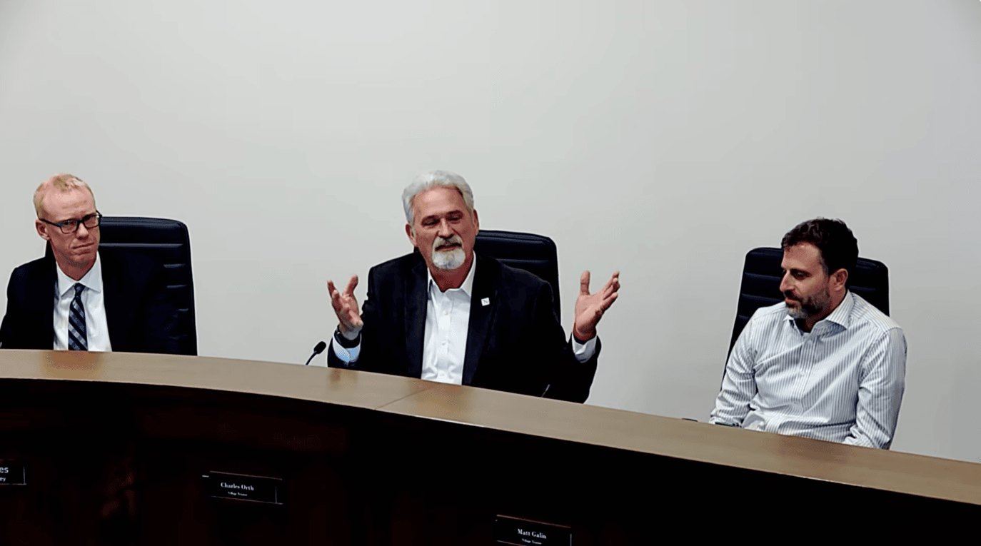 Three men seated at a board meeting dais, the center one speaking and gesturing with both hands.