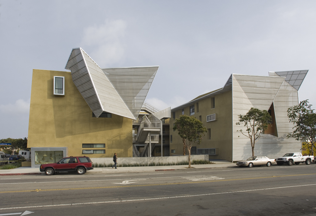 Multi-story affordable housing building with yellow stucco exterior and angular metallic roof elements on a city street.