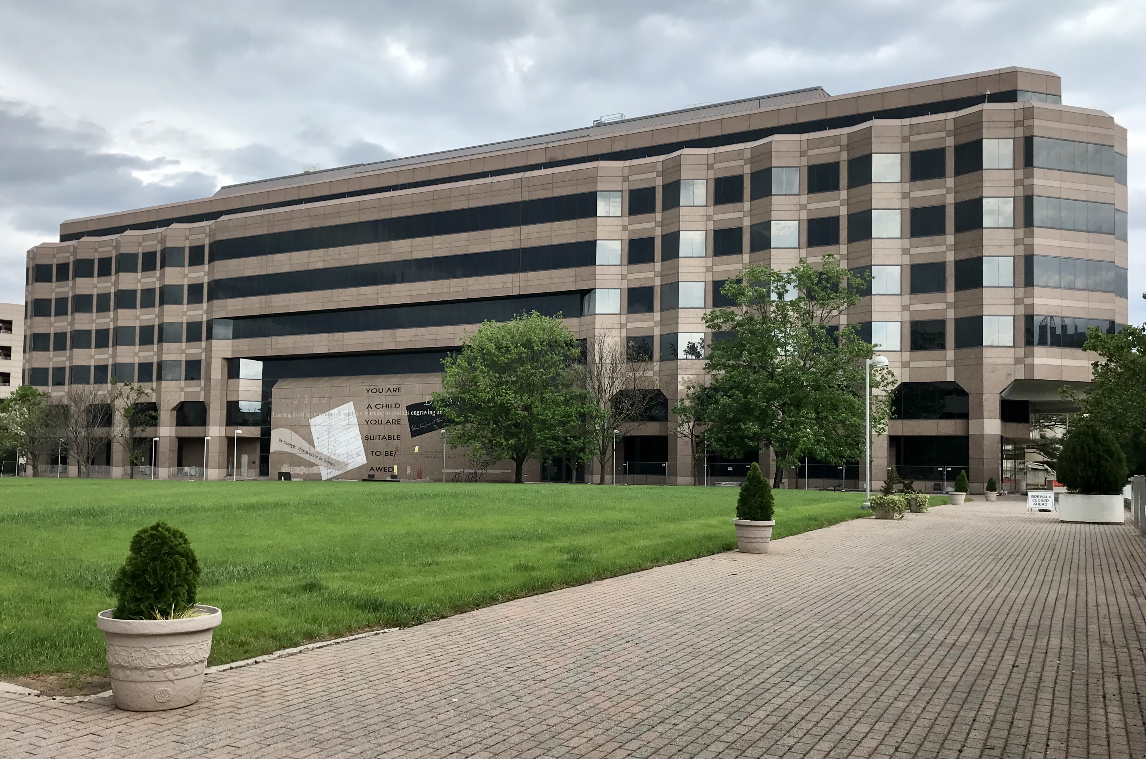 Multi-story tan office building with brick plaza and manicured lawn under an overcast sky.