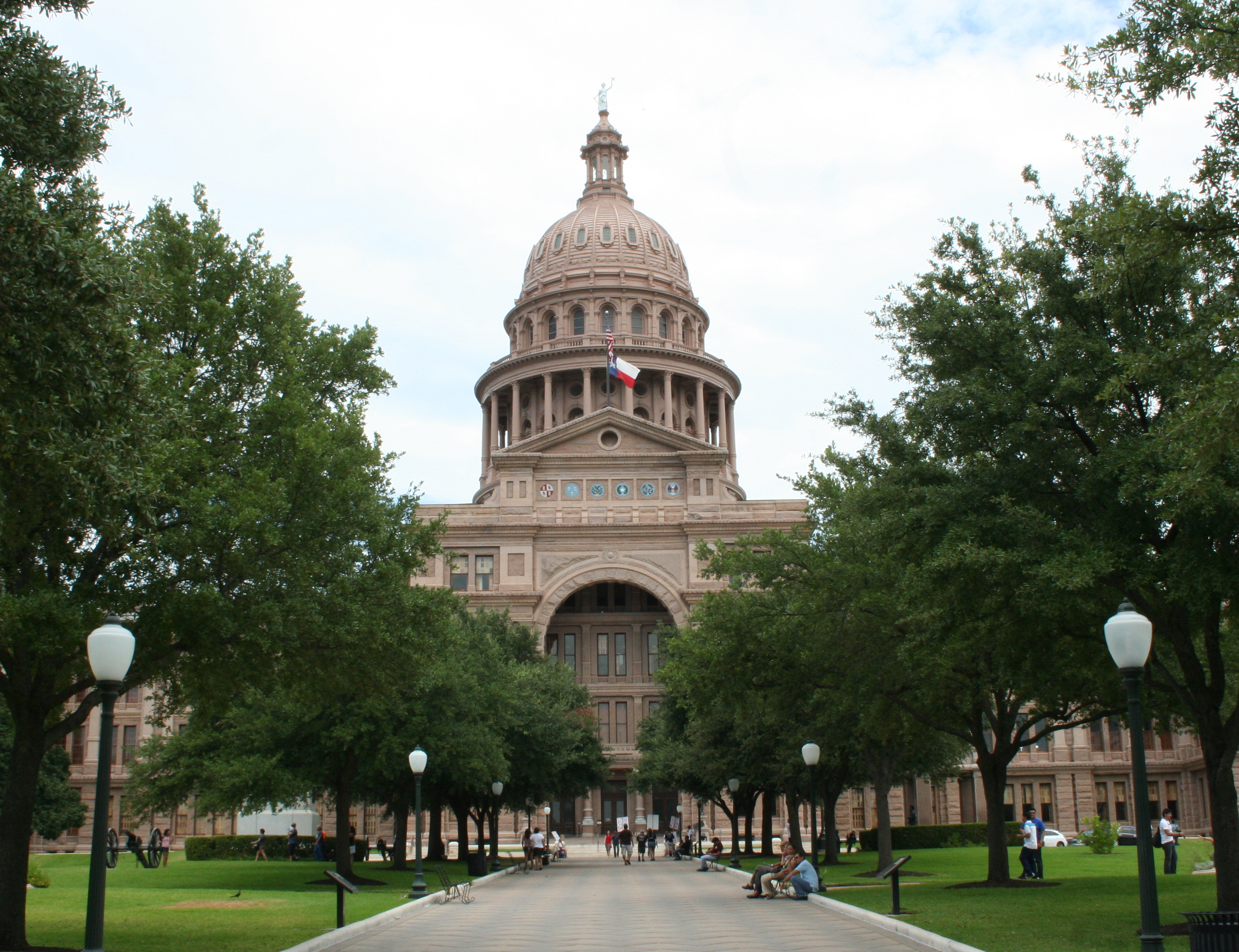 The Texas State Capitol building seen from the main walkway, flanked by trees and lamp posts.