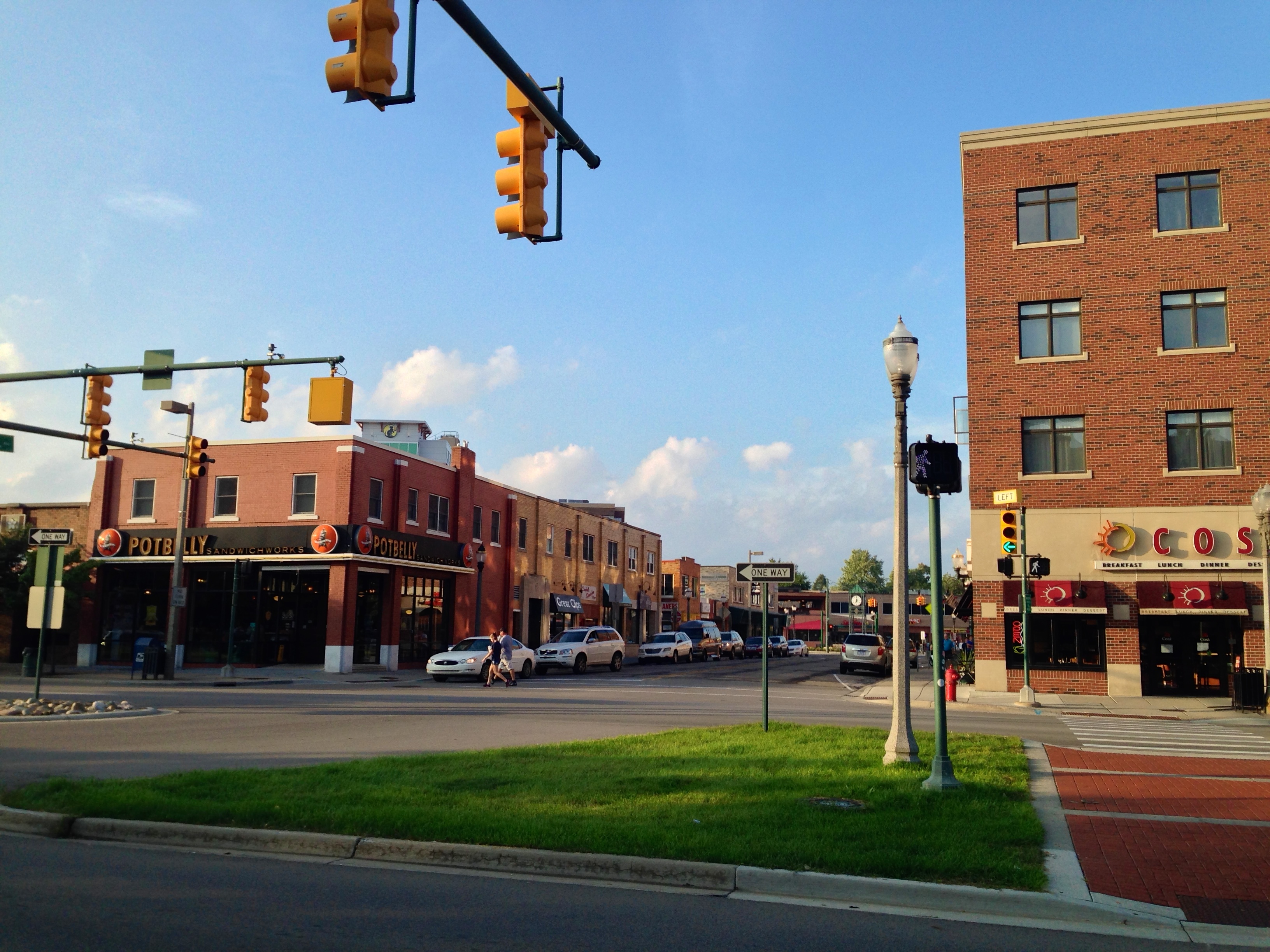 Brick commercial storefronts and traffic signals line a downtown urban avenue on a partly cloudy day.