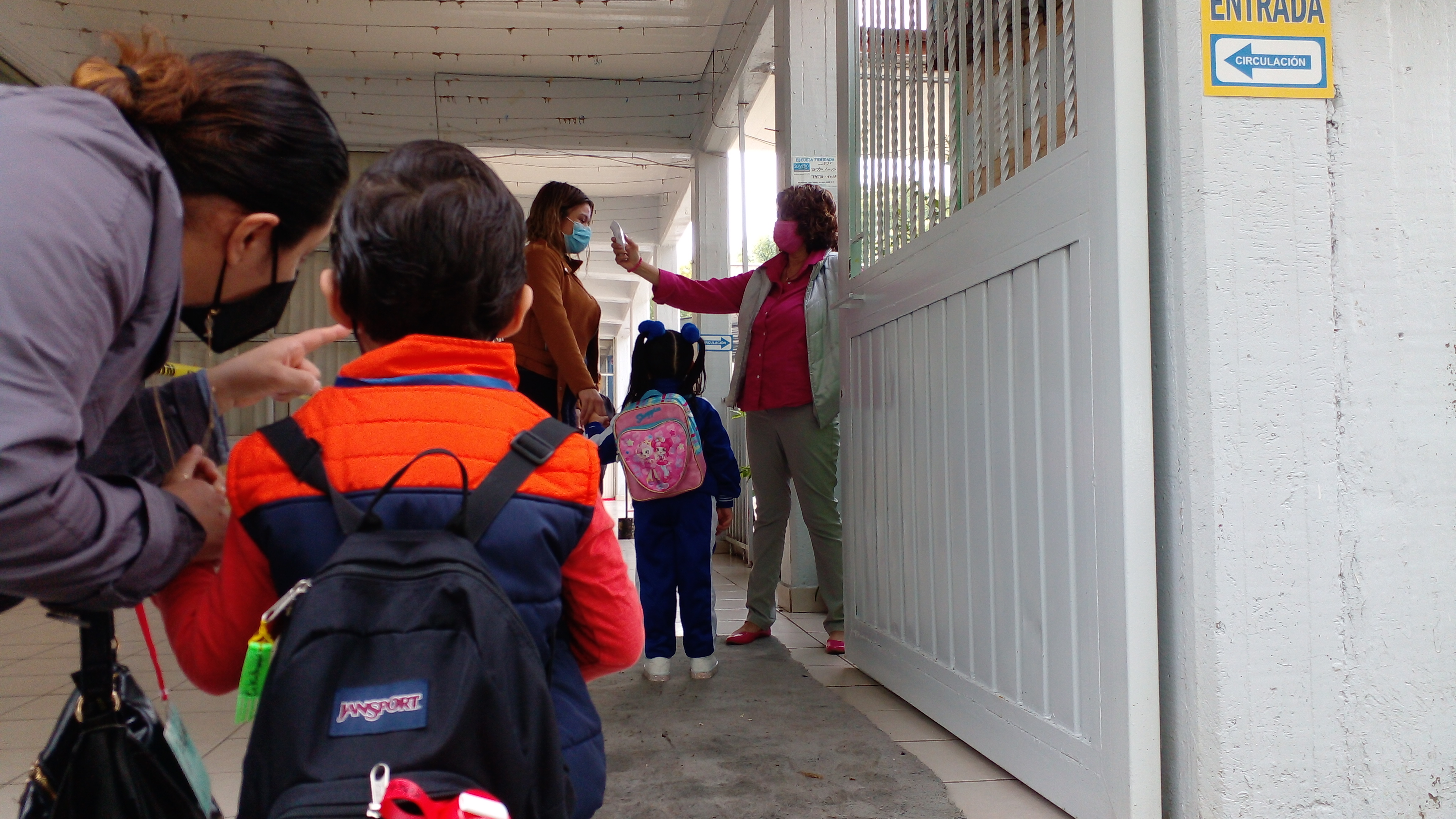 Young children with backpacks lining up at a school entrance gate with adult supervision.