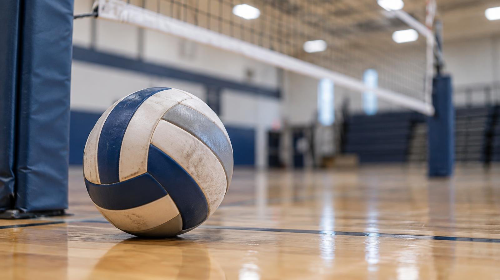 A volleyball resting on a hardwood gymnasium floor near a net post.
