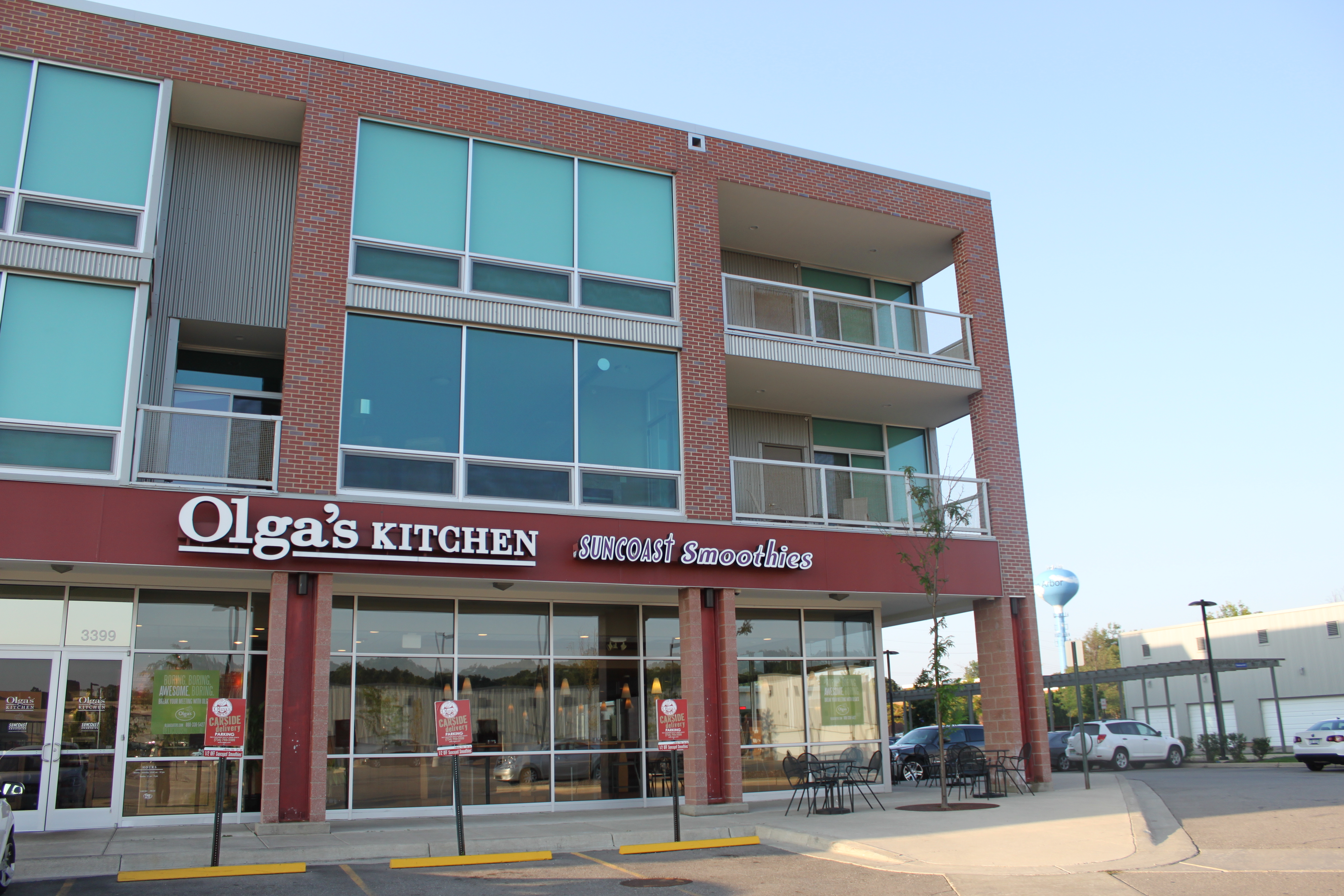 Exterior of an Olga's Kitchen restaurant in a multi-story brick building with outdoor seating.