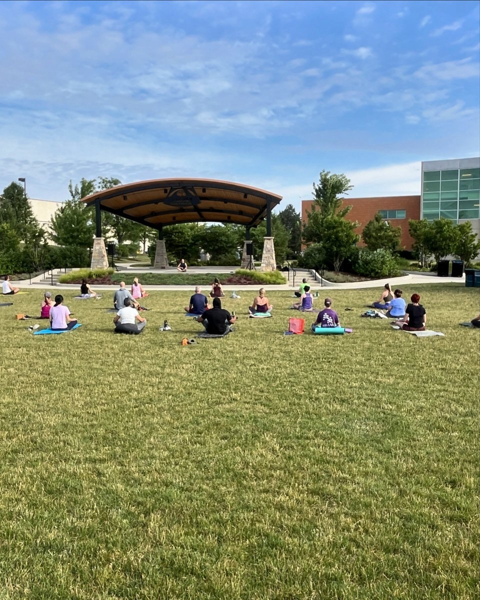People sitting on yoga mats on a green lawn facing a covered pavilion structure on a partly cloudy day.