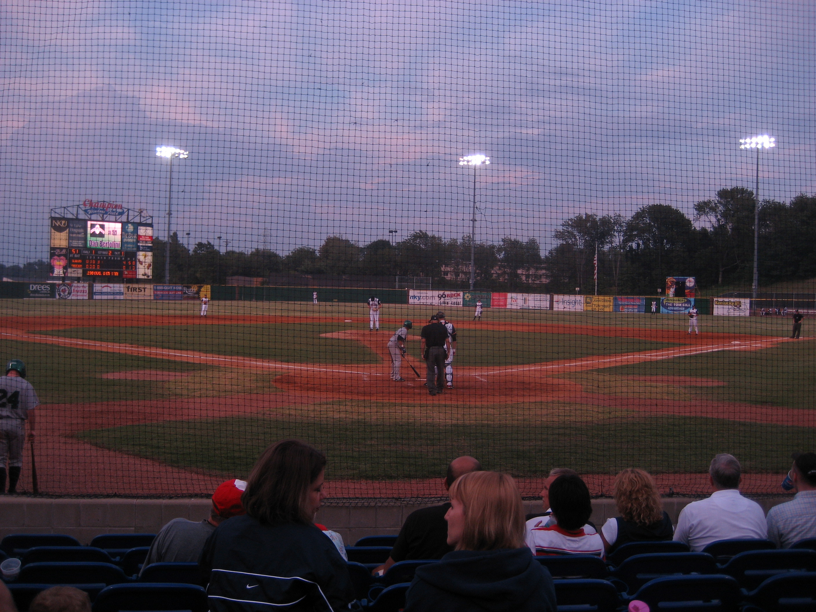 Minor league baseball game viewed from behind home plate netting under stadium lights at dusk.