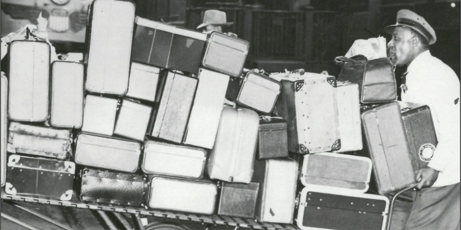 Black-and-white photo of a man in a uniform cap beside a cart piled high with suitcases.