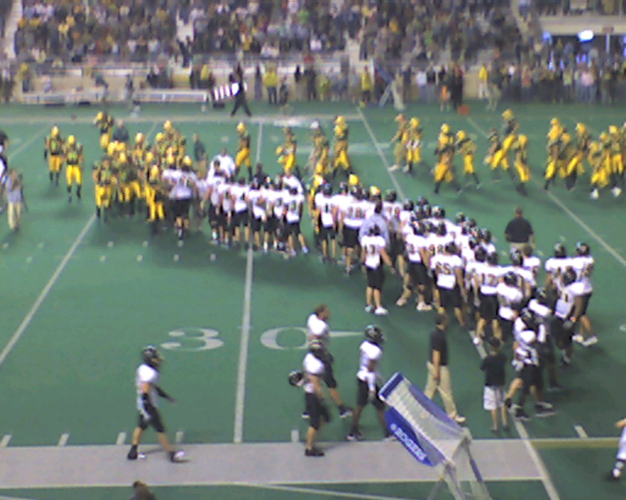 College football players in gold uniforms compete on indoor turf before a large crowd.
