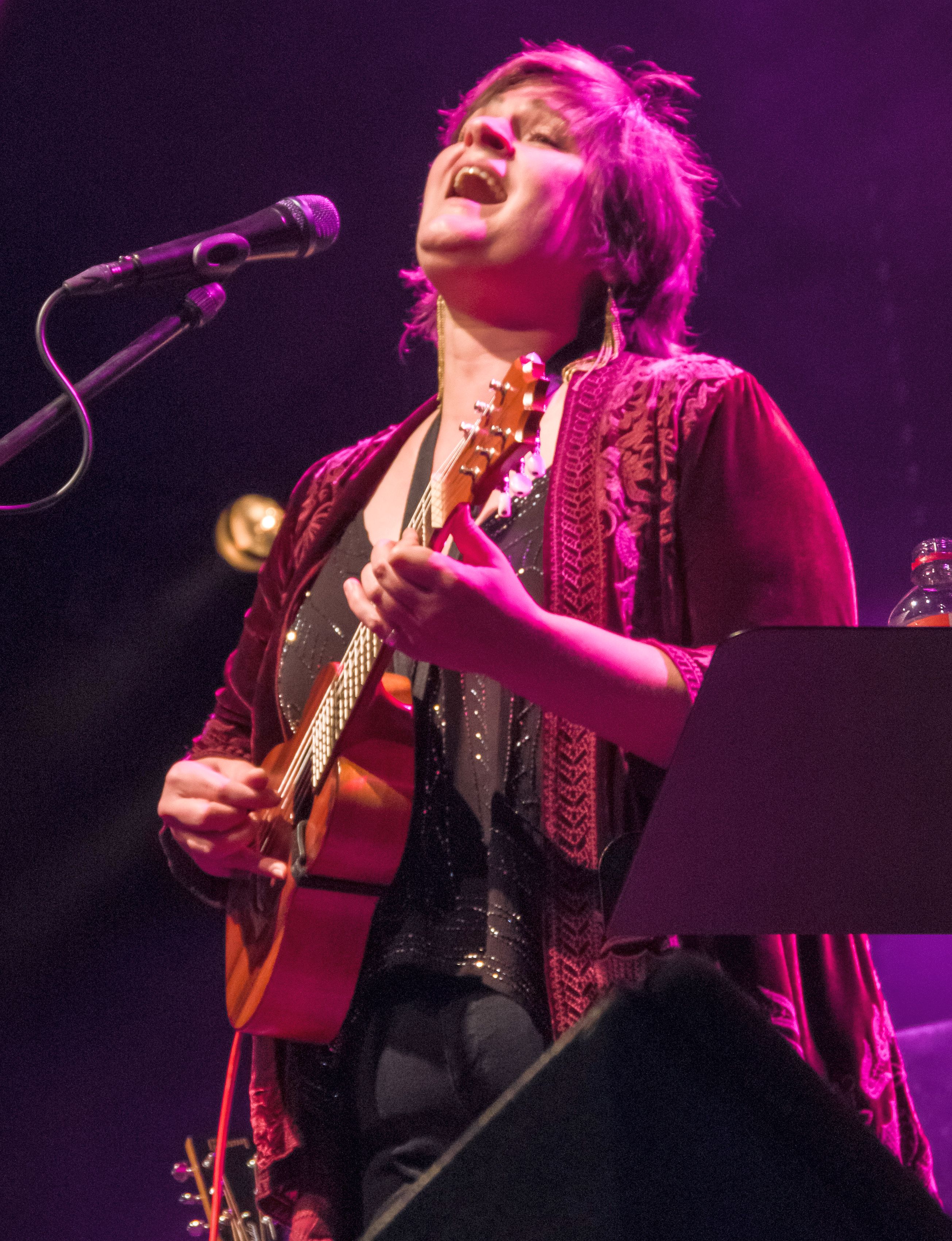 A woman plays acoustic guitar and sings into a microphone under purple stage lighting.