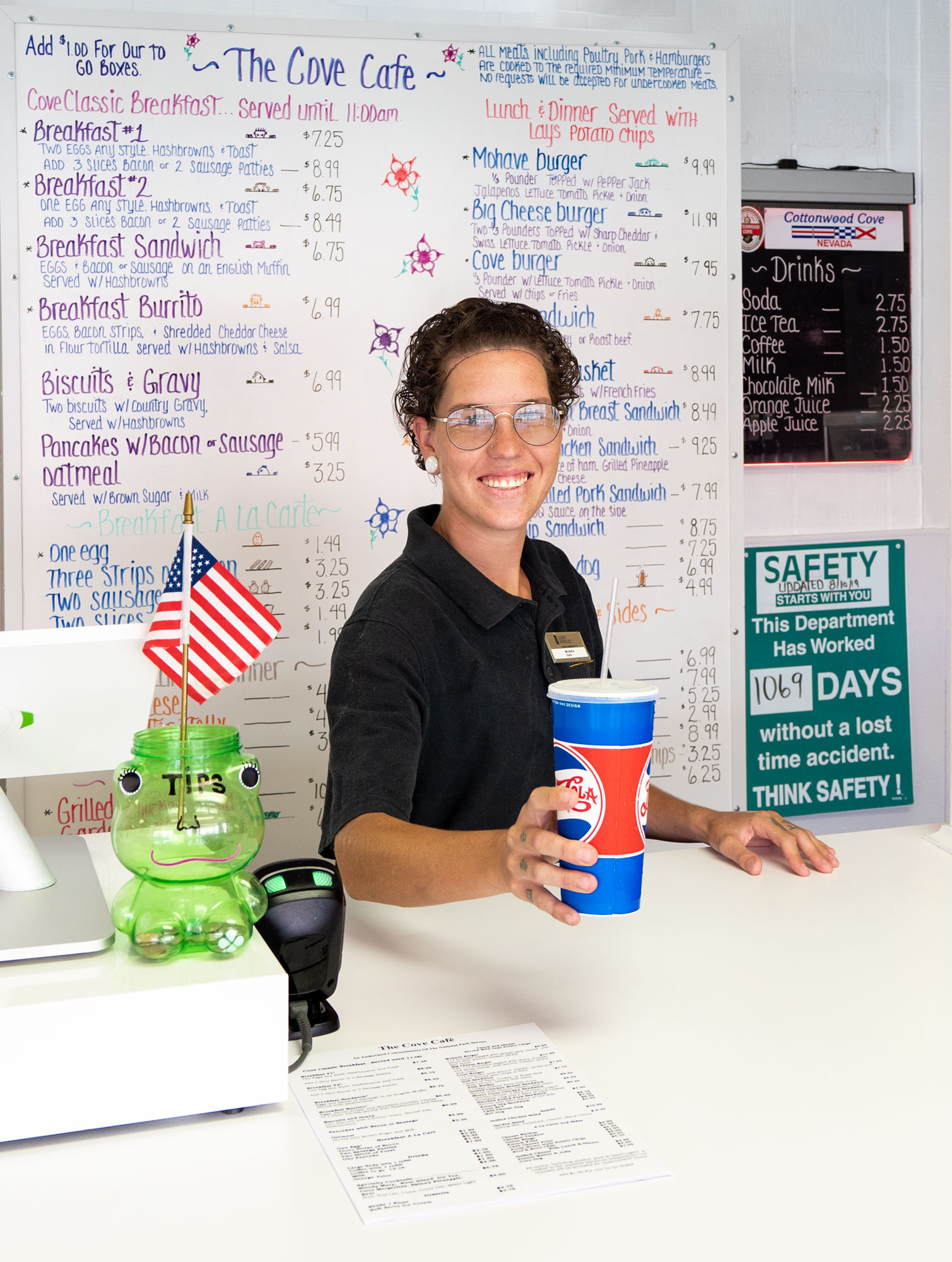 Smiling cafe worker behind counter with a tip jar and menu board.