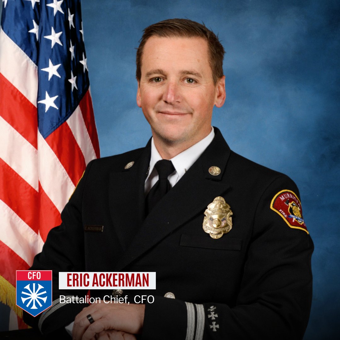 Man in dark fire department dress uniform with gold badge posed before an American flag.