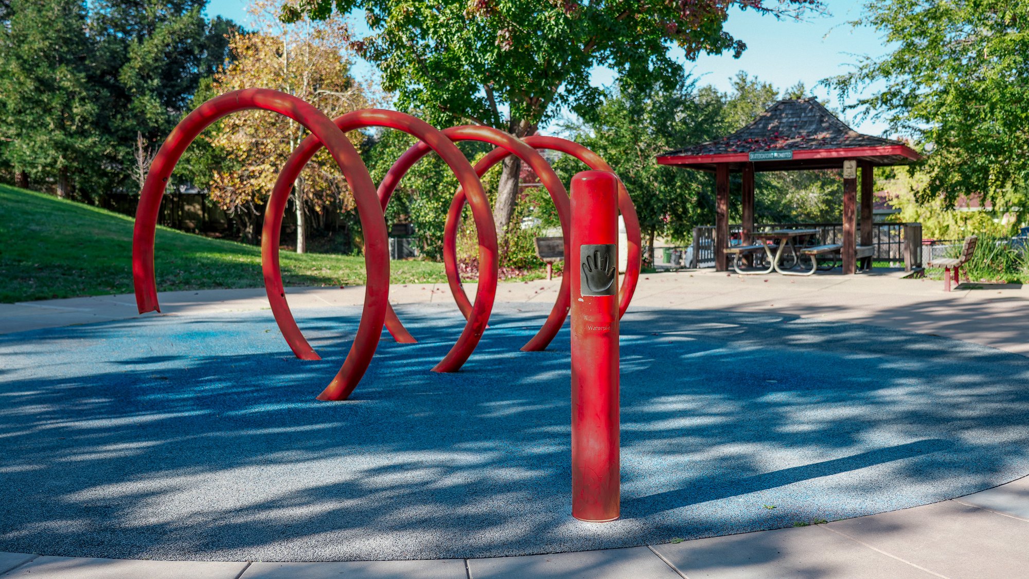 Red arched metal splash pad structures on a blue rubberized surface in a park.