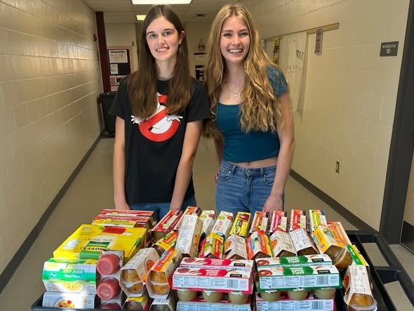 Two students stand behind a cart loaded with fruit cups and applesauce in a school hallway.