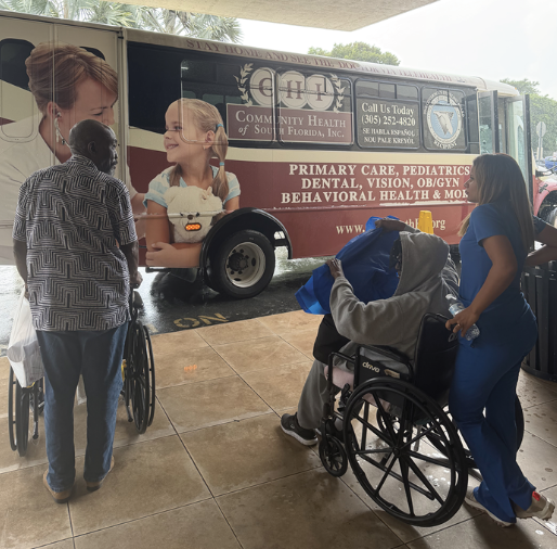 A healthcare worker in blue scrubs assists a wheelchair user near a community health mobile bus.
