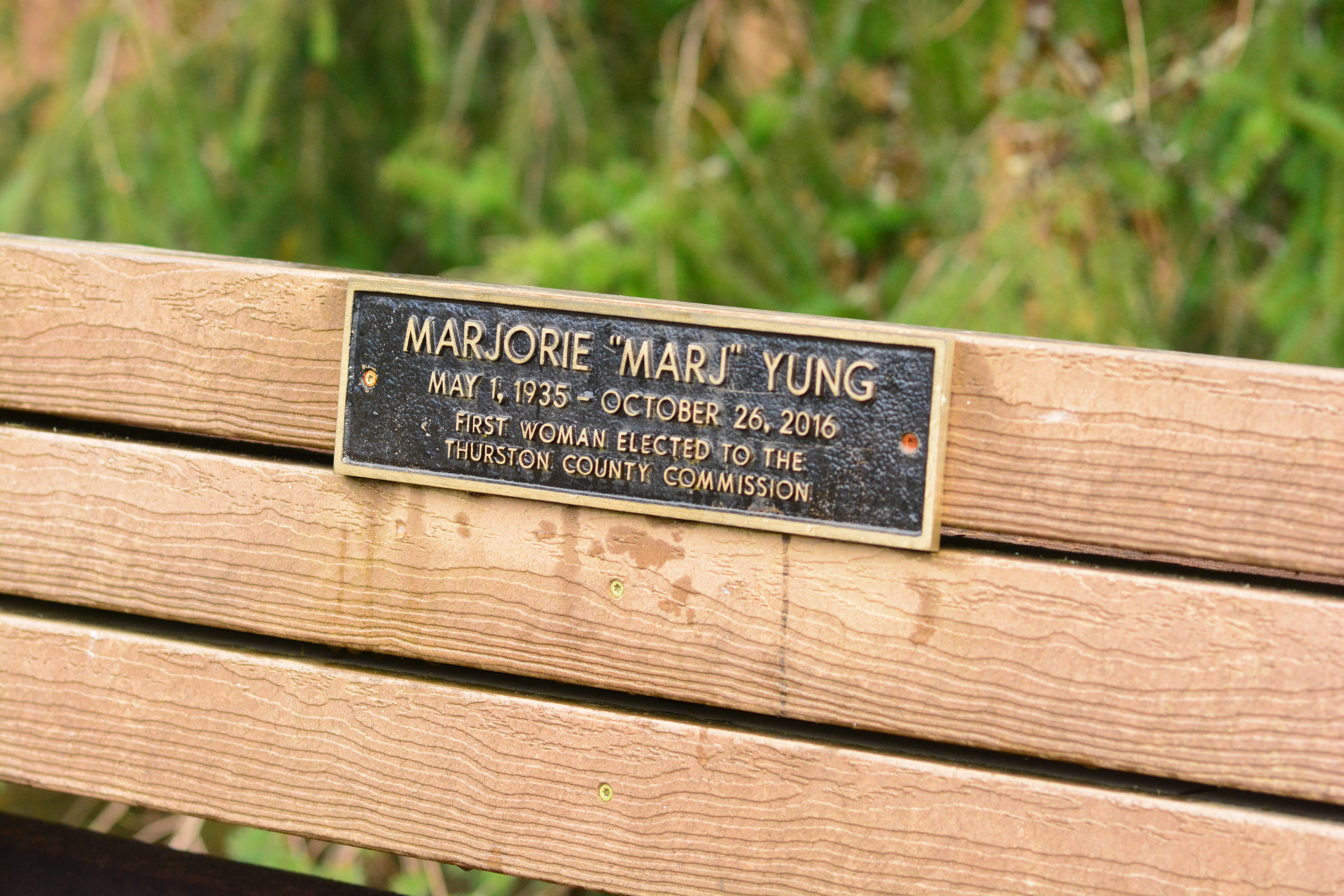 Wooden park bench with a bronze memorial plaque honoring Marjorie Yung.