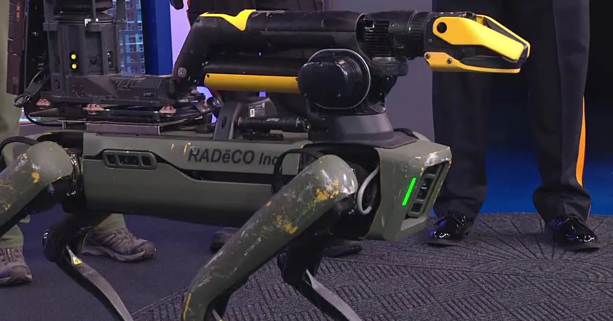 A military-green four-legged robot with a yellow and black sensor arm mounted on top.