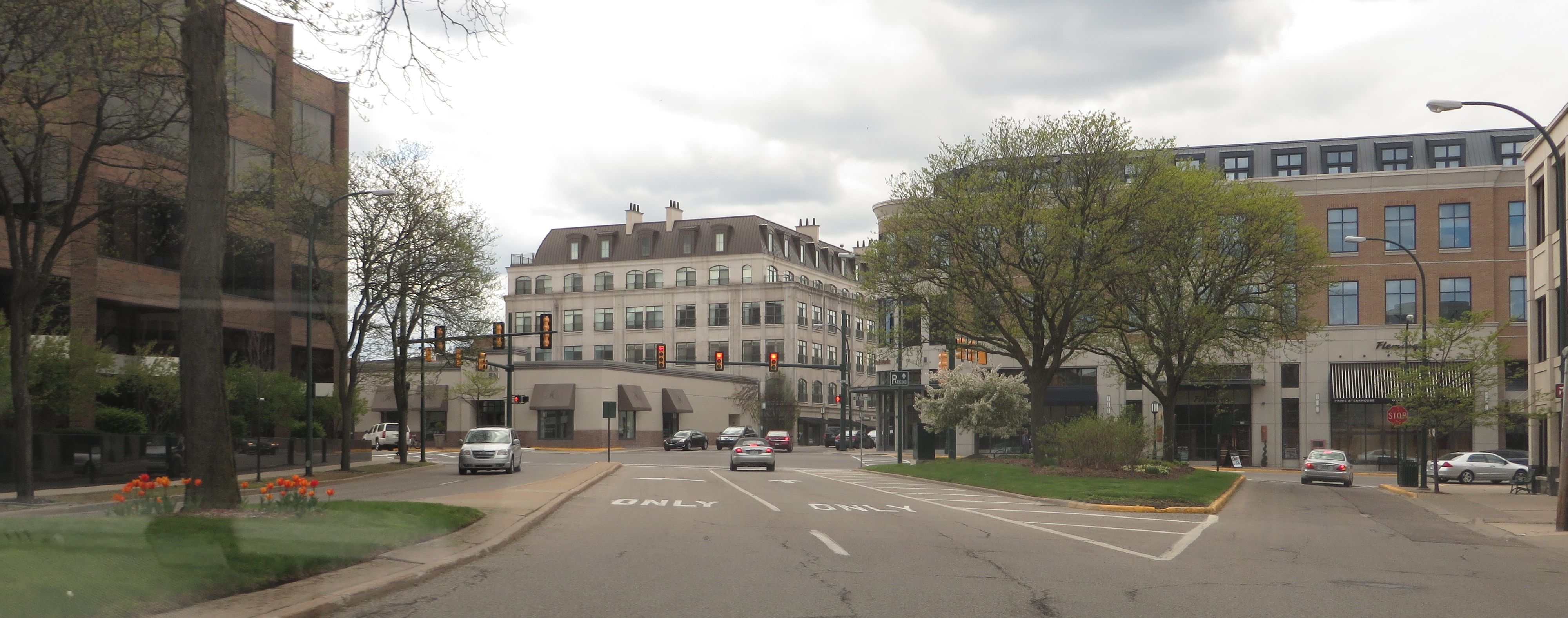 Downtown street intersection with commercial buildings, trees, and traffic signals under a cloudy sky.