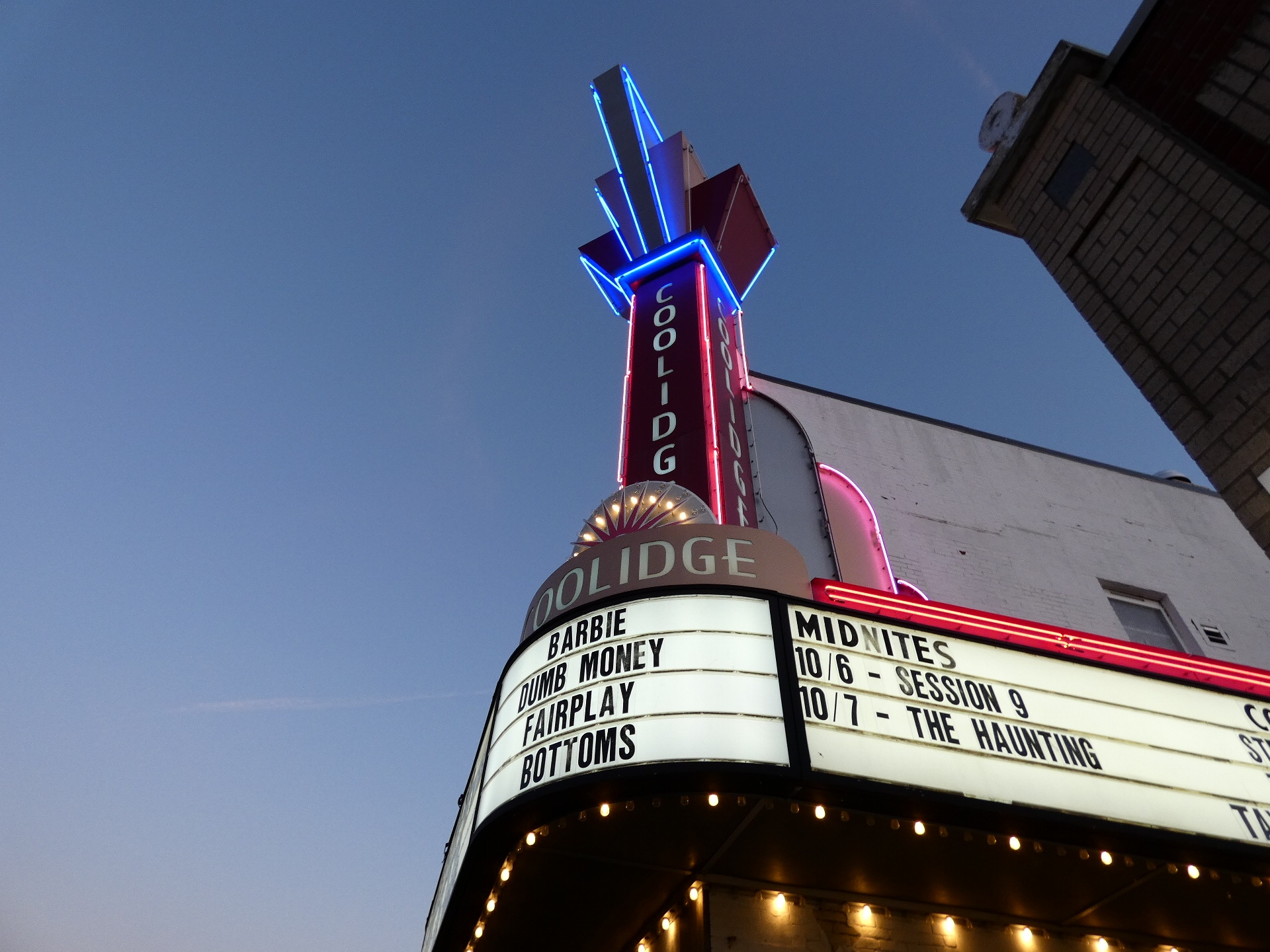 Exterior marquee and neon tower of a movie theater at dusk with film titles displayed.