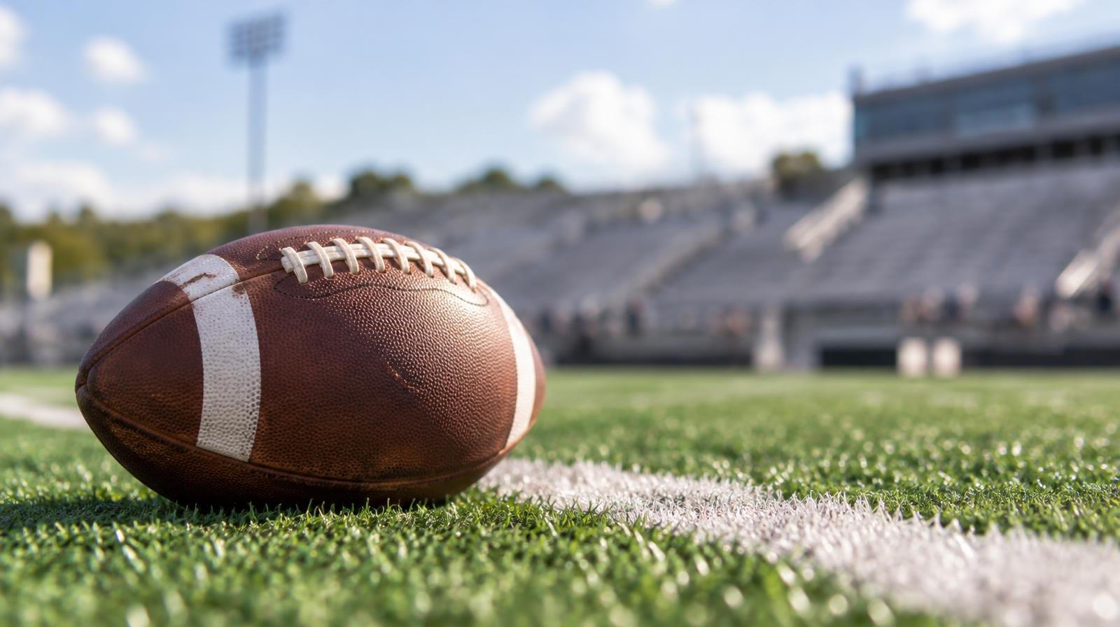 A football resting on green turf beside a painted white yard line, stadium bleachers blurred behind it.