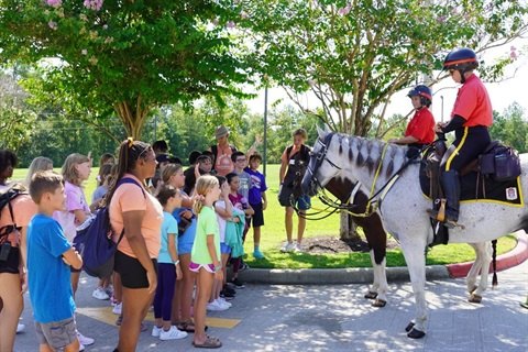 Two uniformed officers on horseback face a crowd of children and adults on a sunny sidewalk.