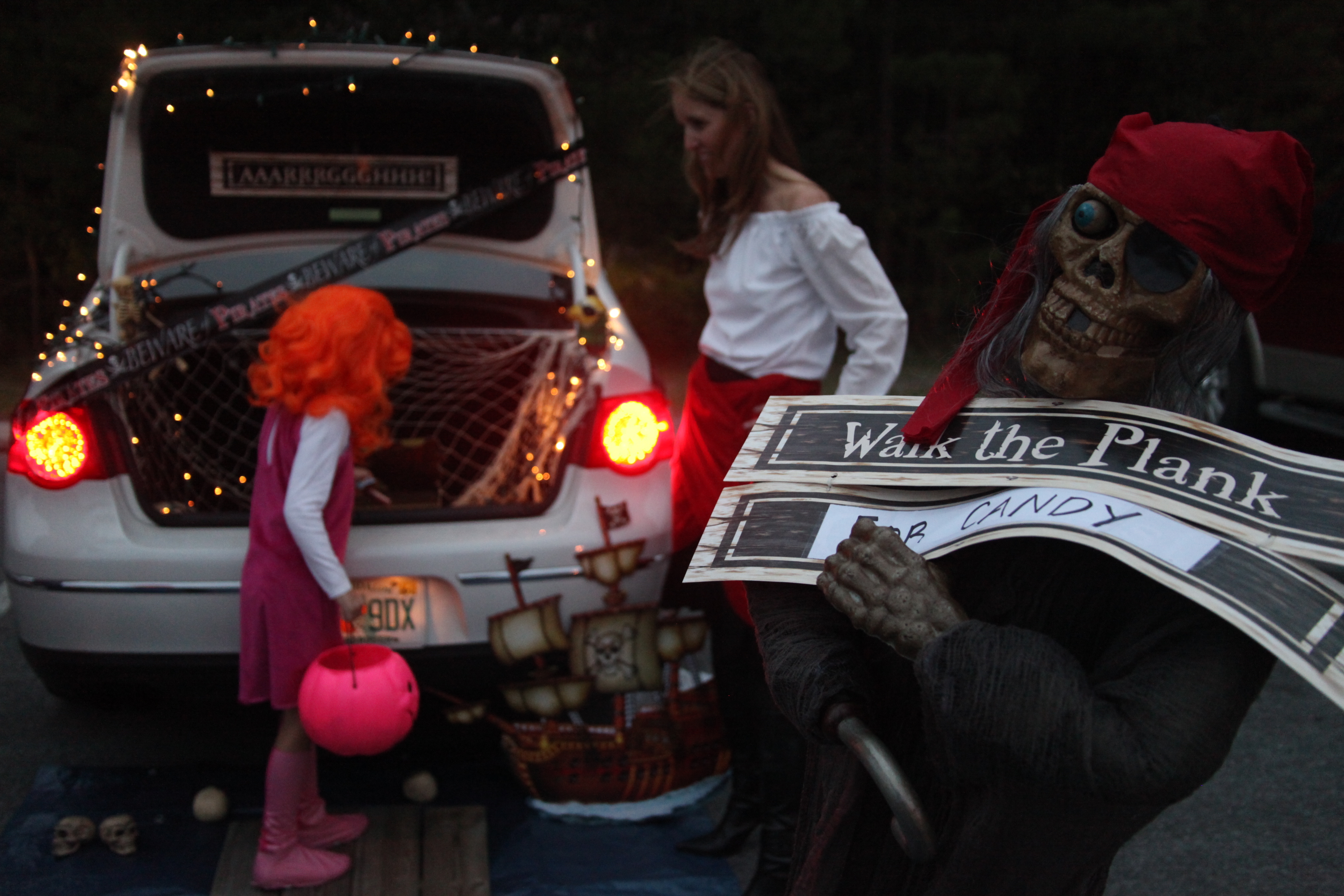 A pirate-themed decorated car trunk with a child in a pink costume holding a pumpkin candy bucket.