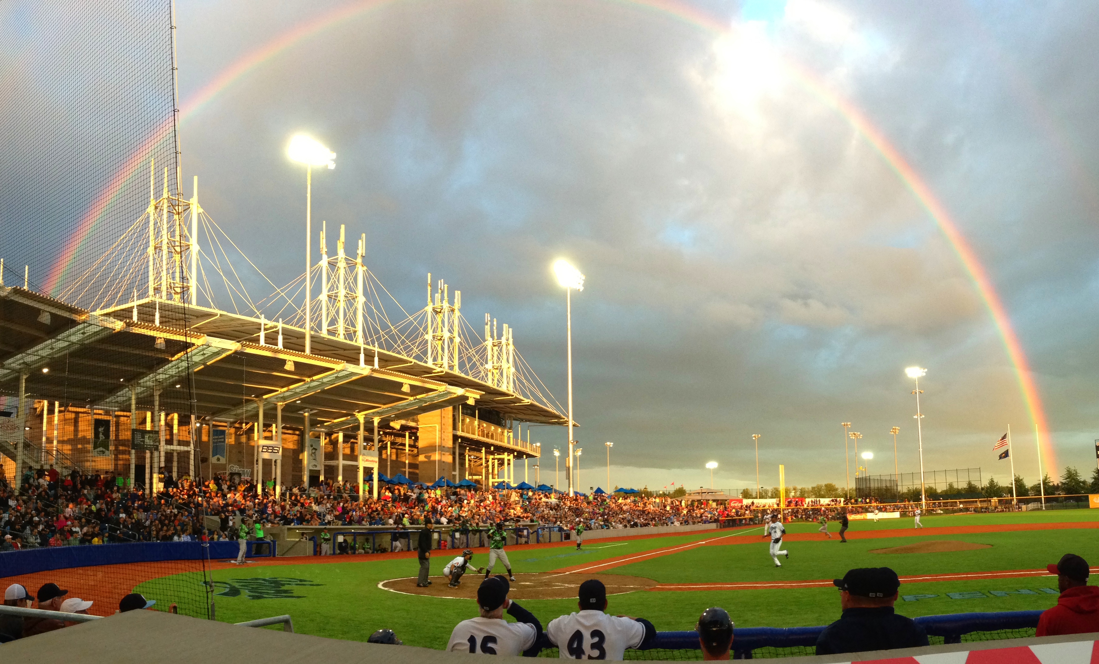 A baseball stadium at night with a full rainbow arching over the grandstand and stadium lights on.