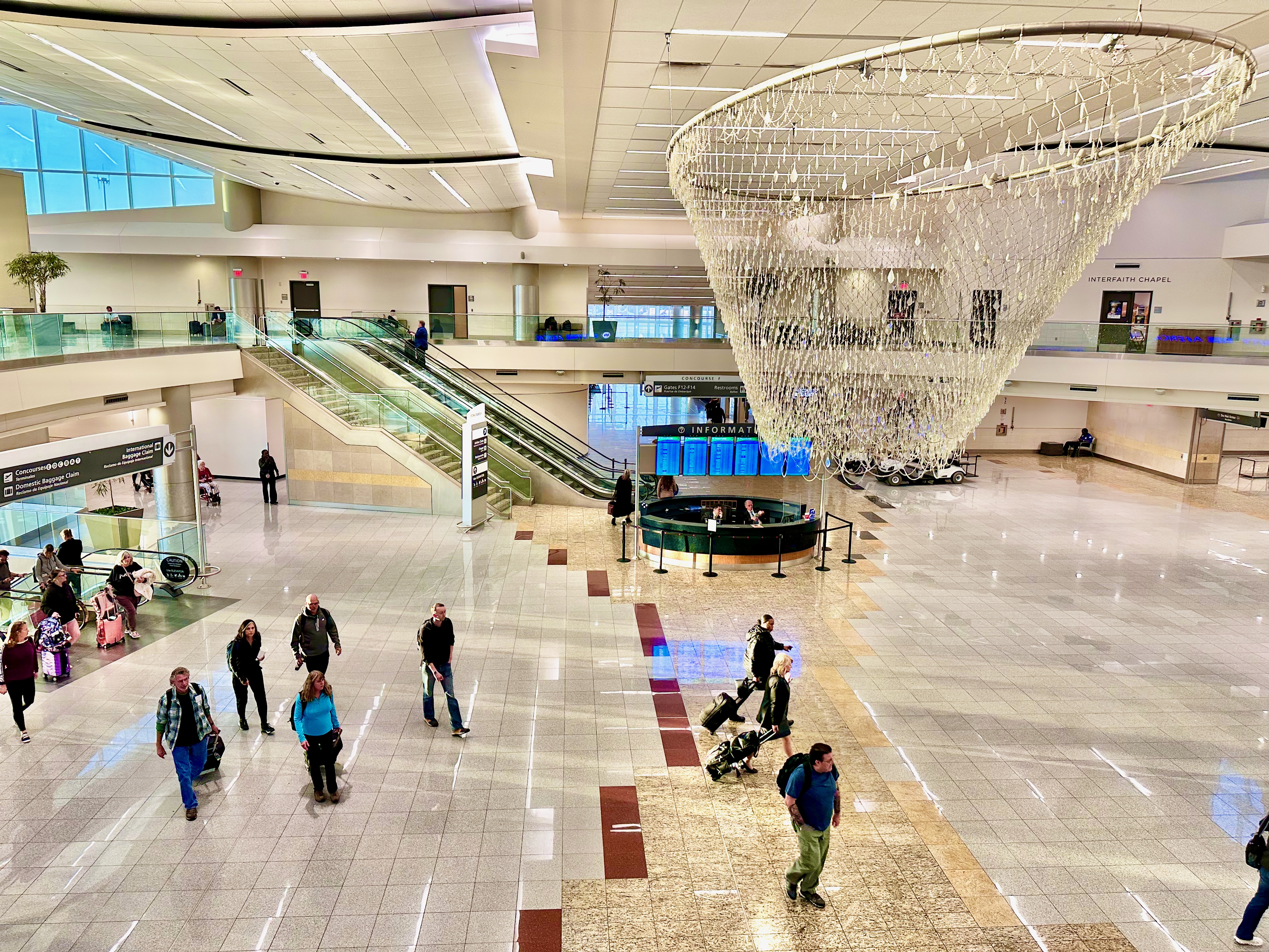 Travelers walk through a large, modern airport terminal with a chandelier and escalators.