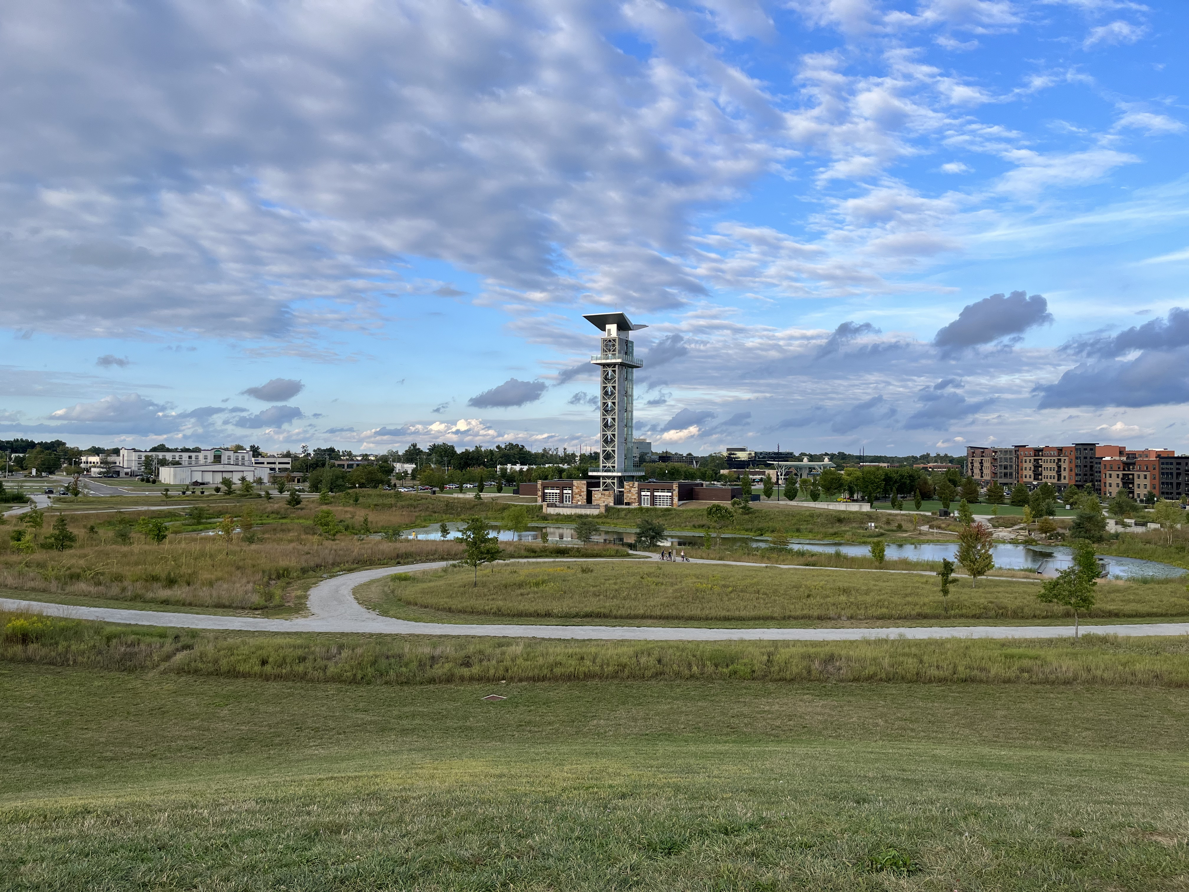 Wide aerial view of a park with an observation tower, pond, and green lawns.
