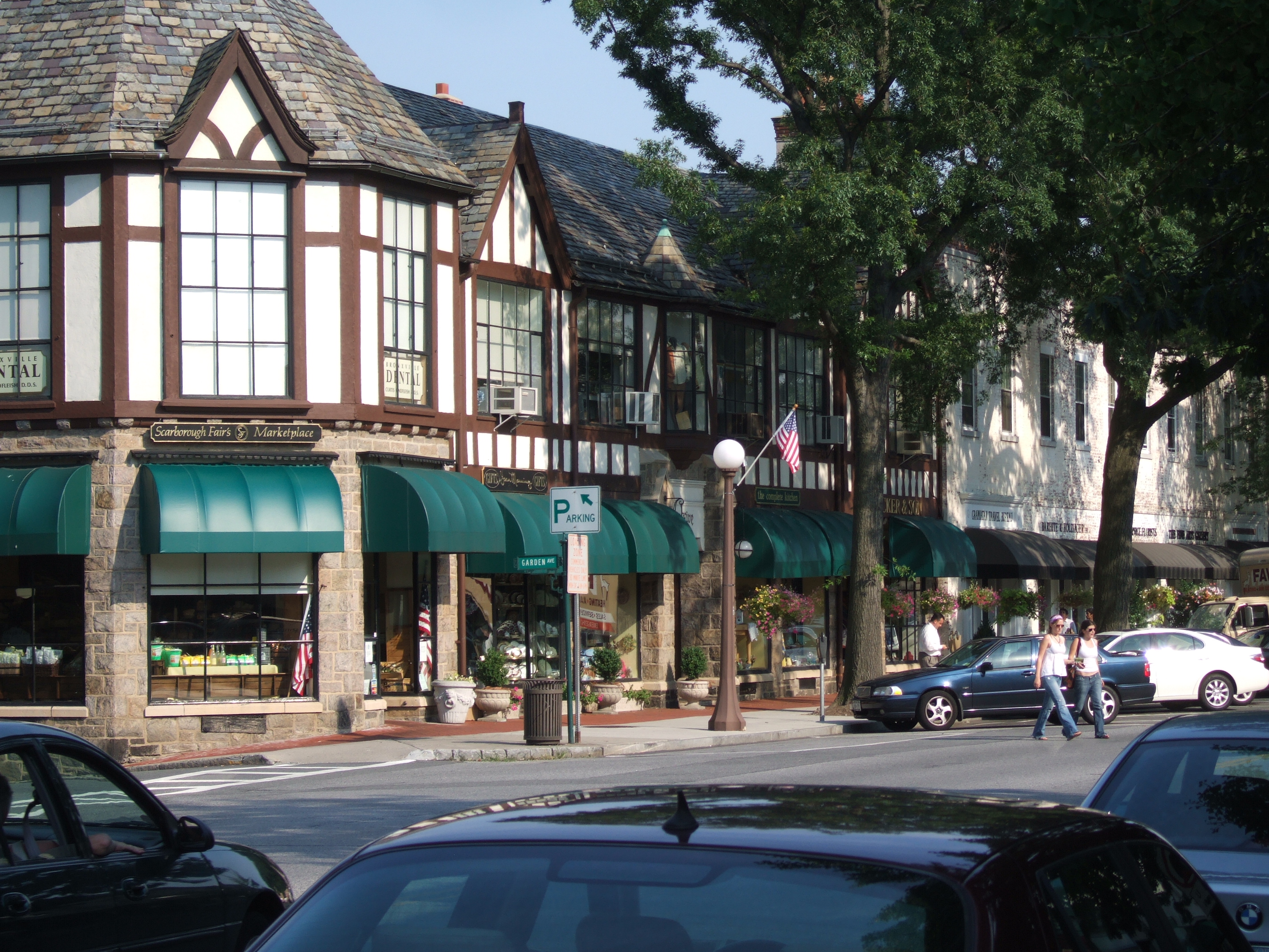Tudor-style shops with green awnings line a tree-shaded village street with parked cars and pedestrians.
