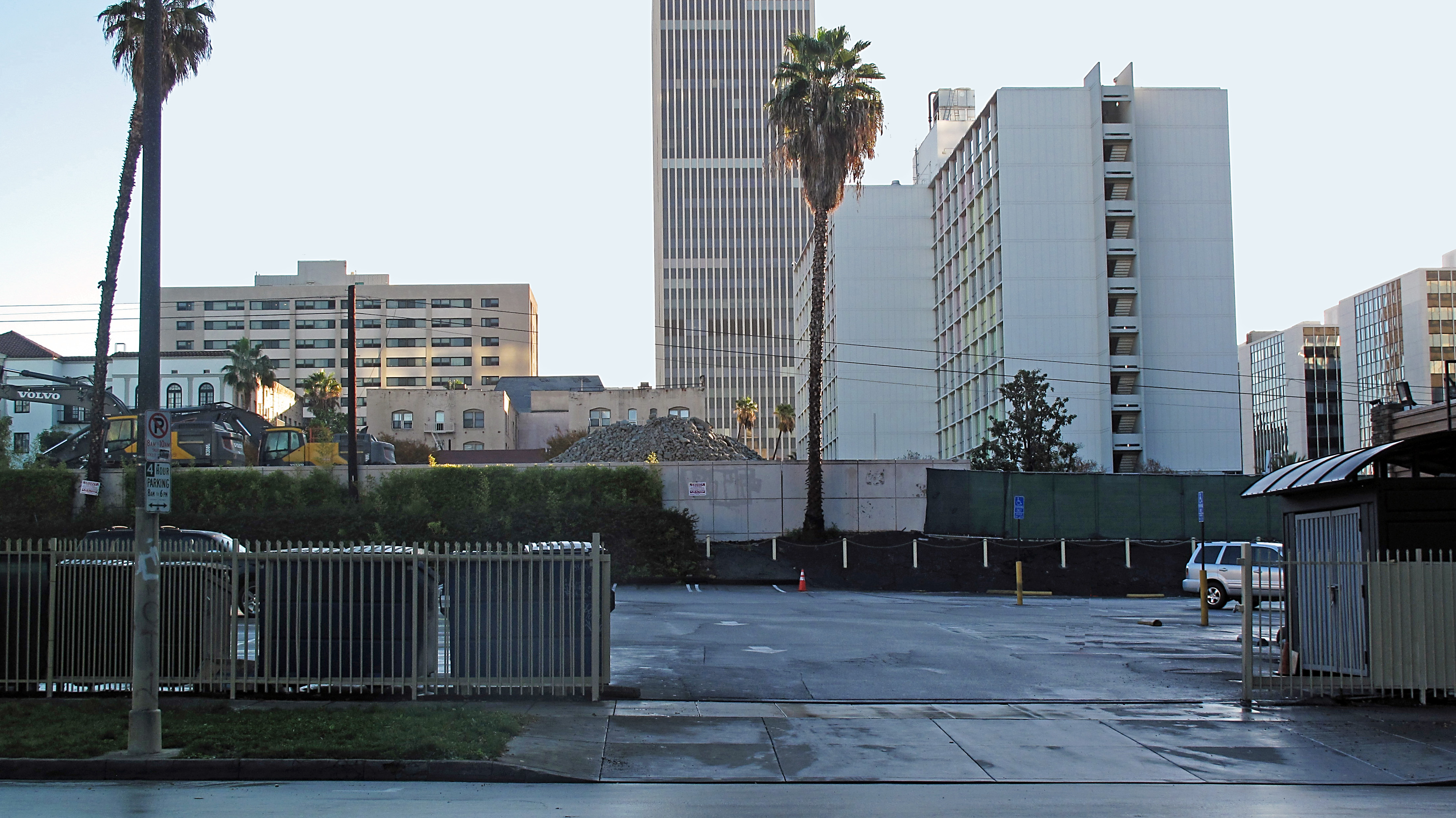 Empty parking lot with demolished building site and office towers behind a metal fence.