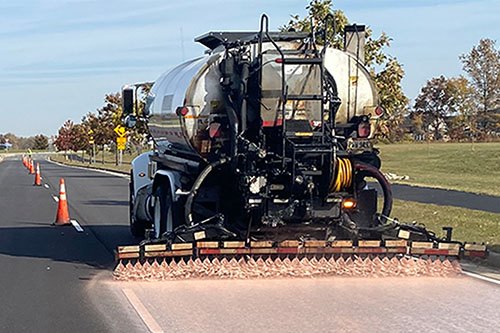 Tanker truck with spray bar applying liquid treatment to a paved road surface.