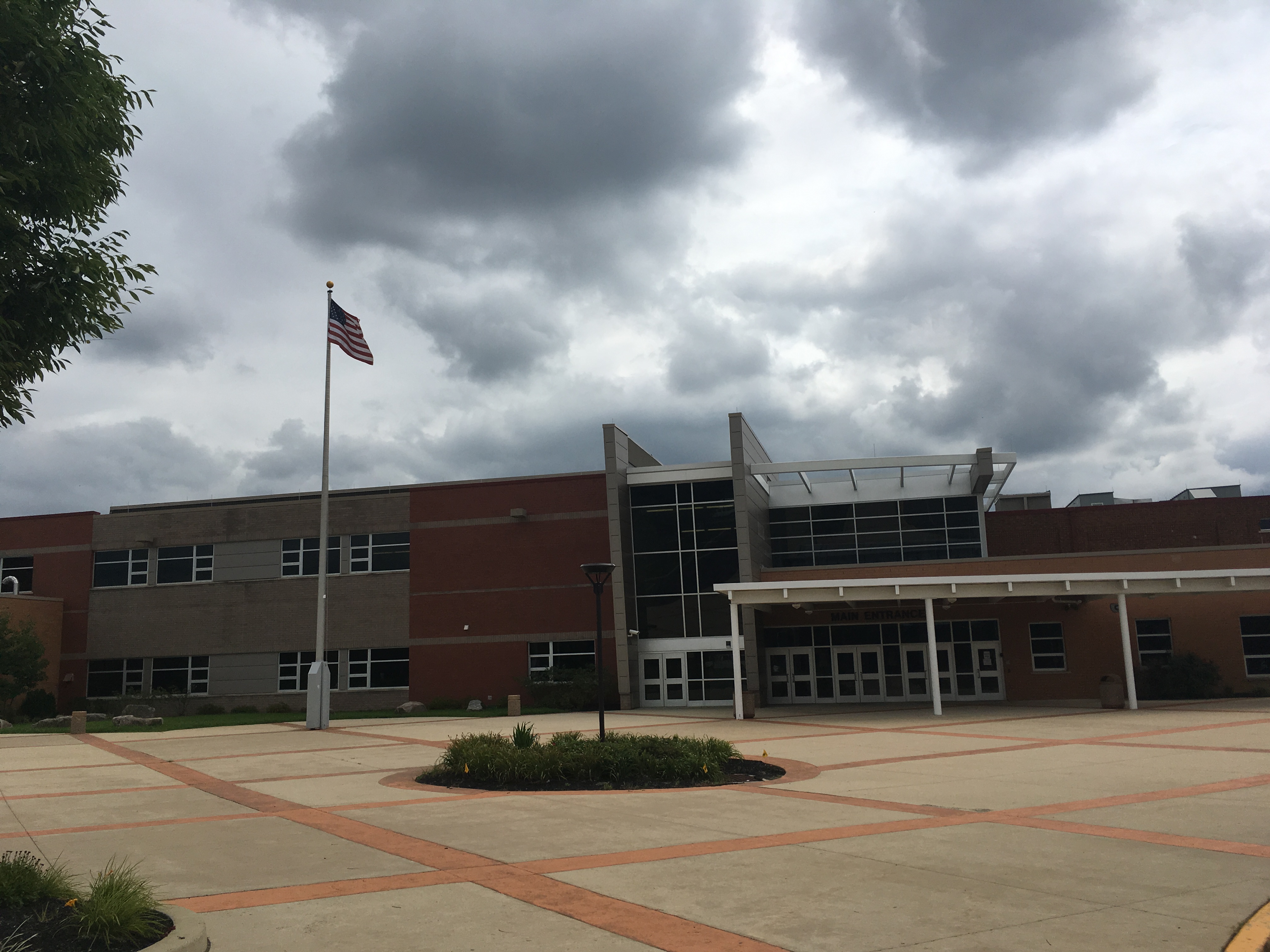 Brick high school building exterior with glass-canopied entrance and American flag on a flagpole.