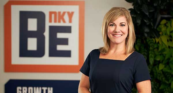 A smiling woman in a navy blazer stands in front of a branded organizational backdrop.