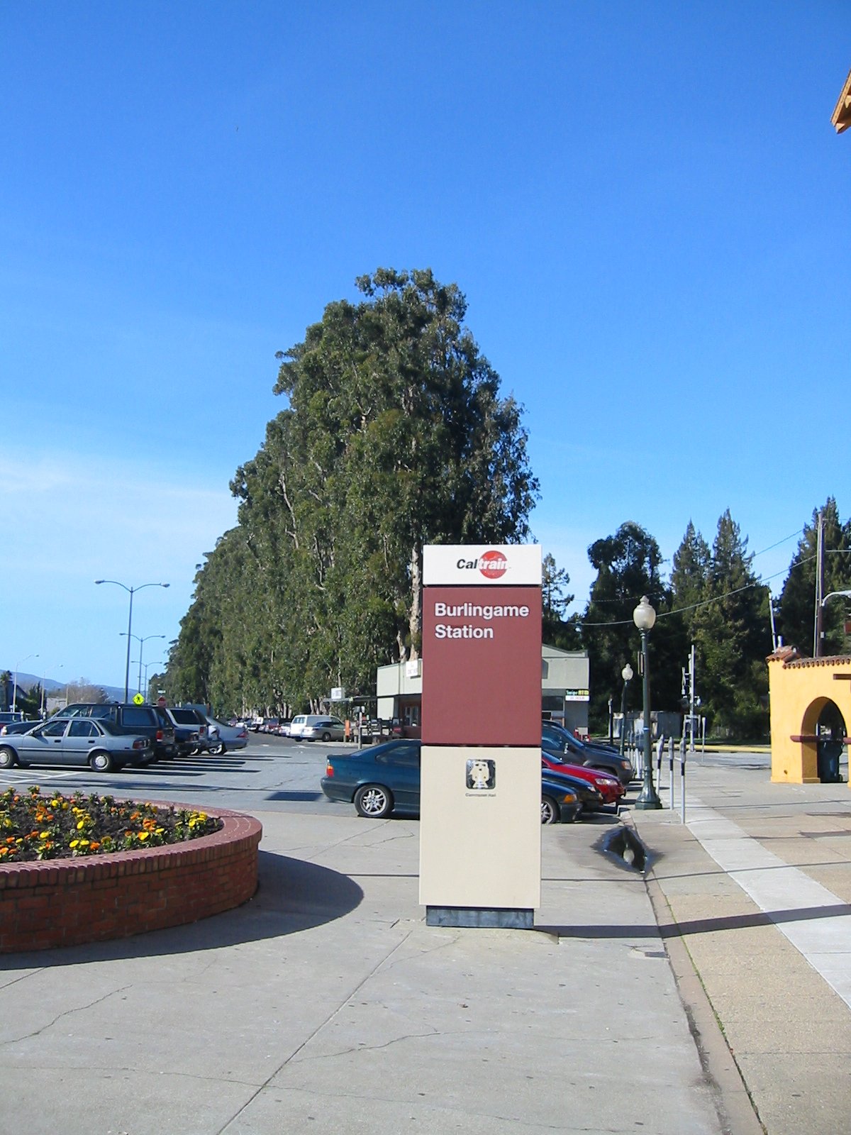 Caltrain station sign reading 'Burlingame Station' with trees and a flower bed nearby.