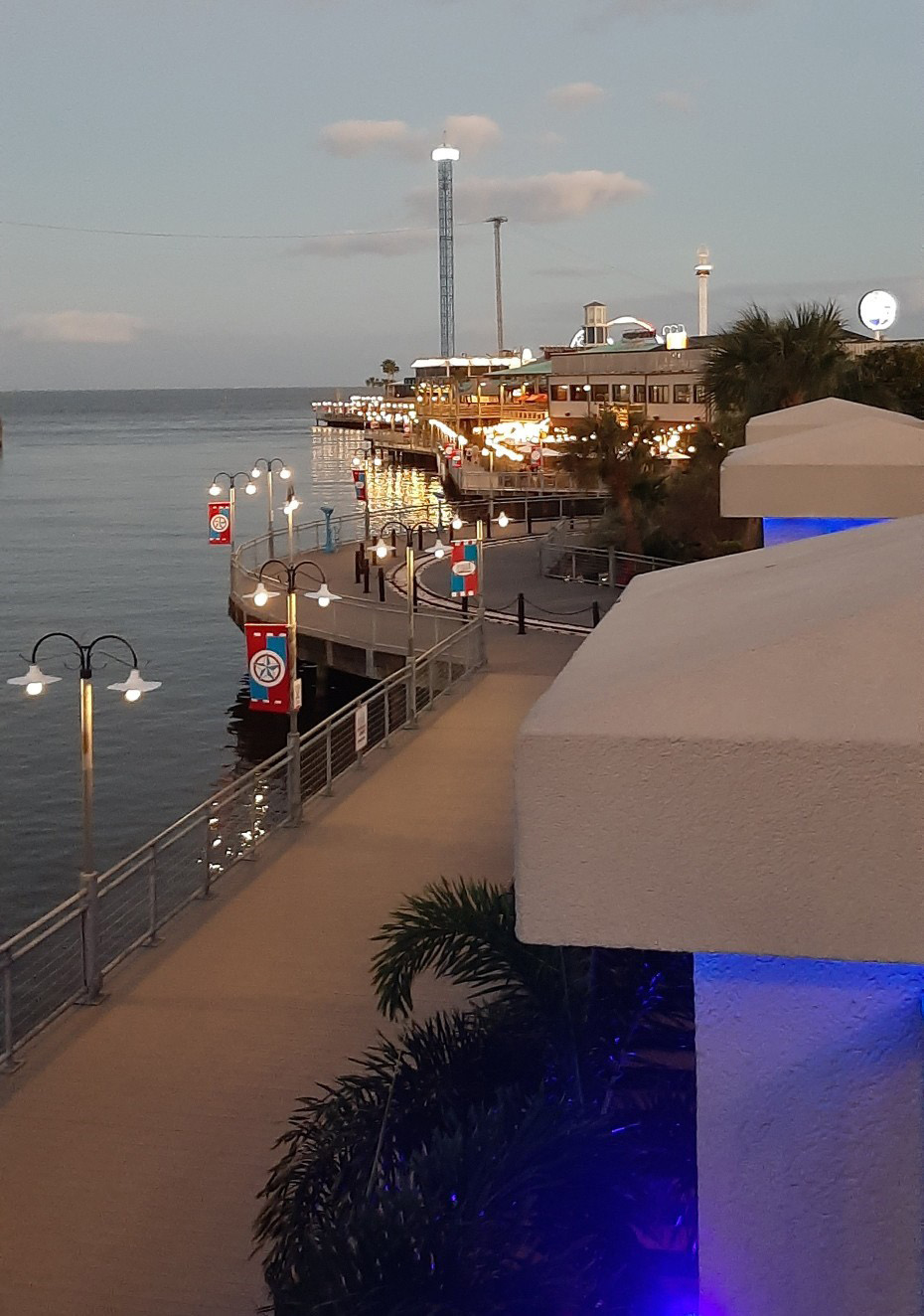 Waterfront boardwalk at dusk with lit restaurants, a drop tower ride, and palm trees.