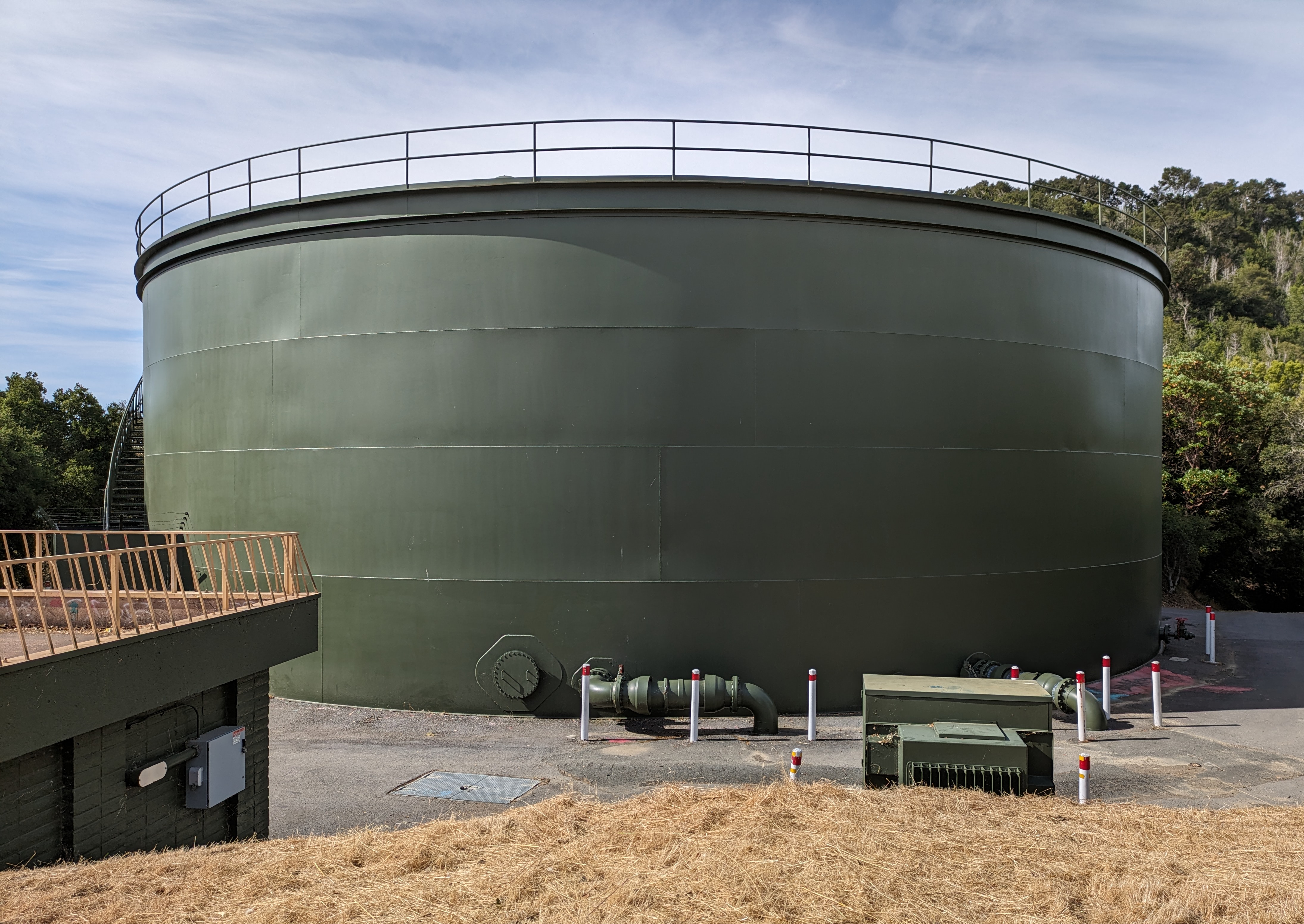 Large dark green cylindrical water storage tank with safety railing and pipe connections at its base.