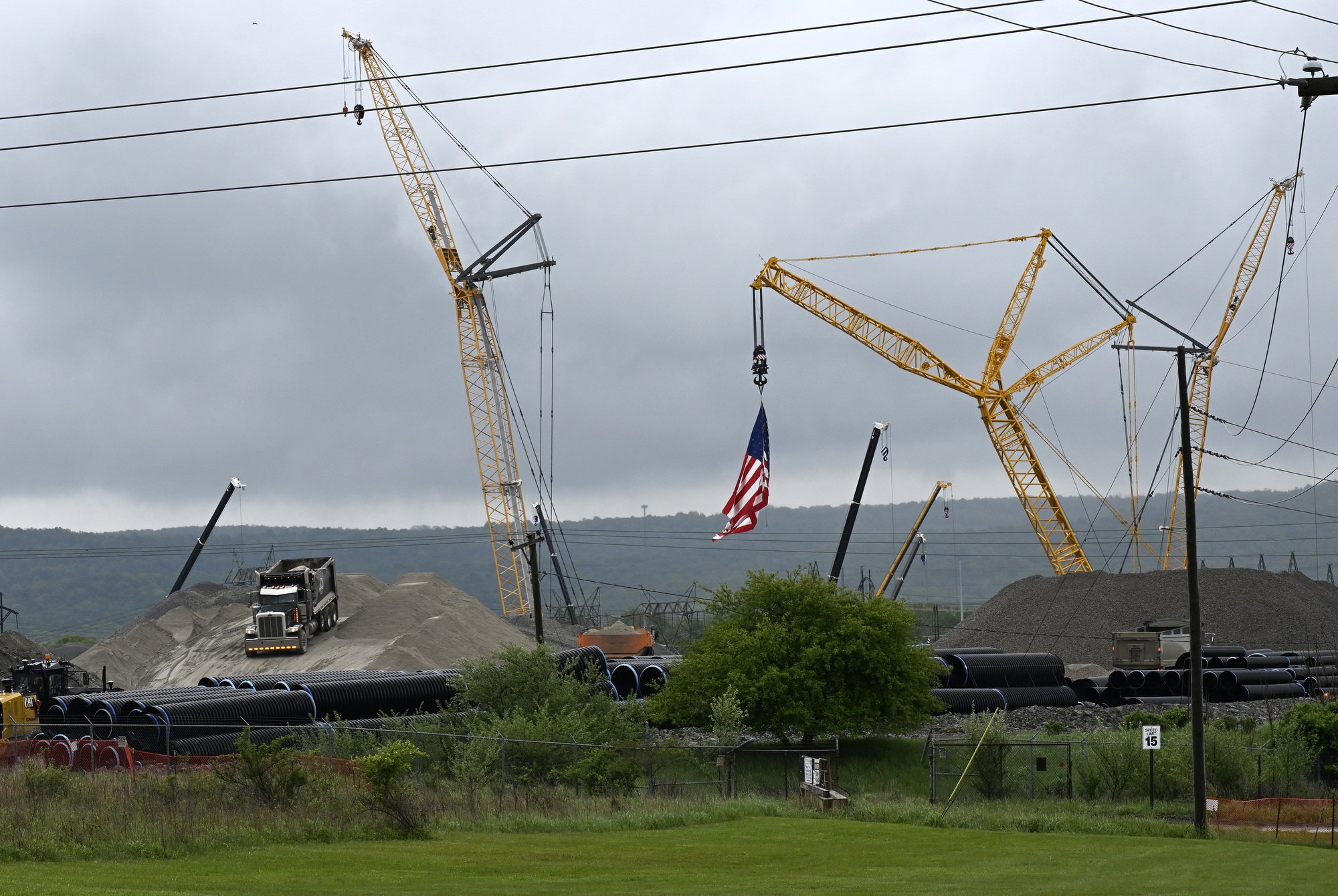 Large construction site with multiple cranes, pipes, gravel piles, and power lines under an overcast sky.