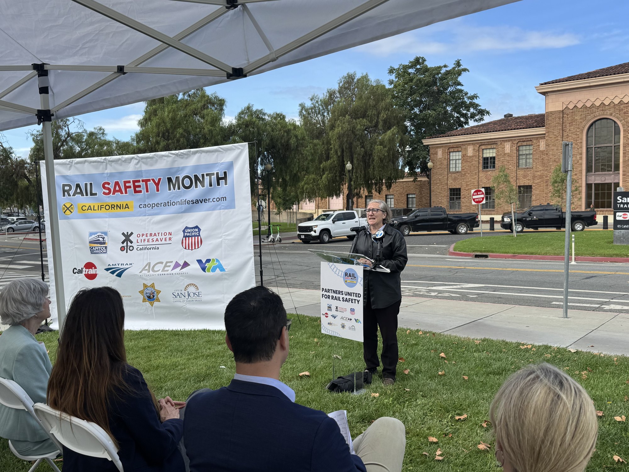 Speaker at outdoor podium under tent with Rail Safety Month California banner.