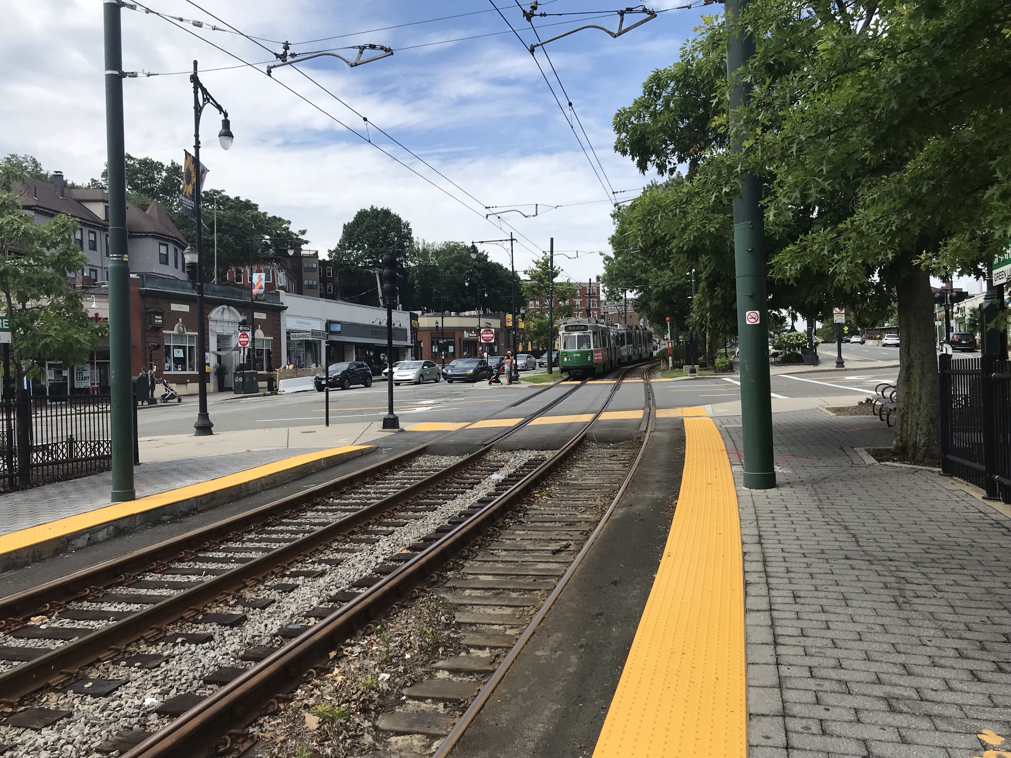 An MBTA Green Line train approaches a station on Beacon Street with storefronts visible across the street.
