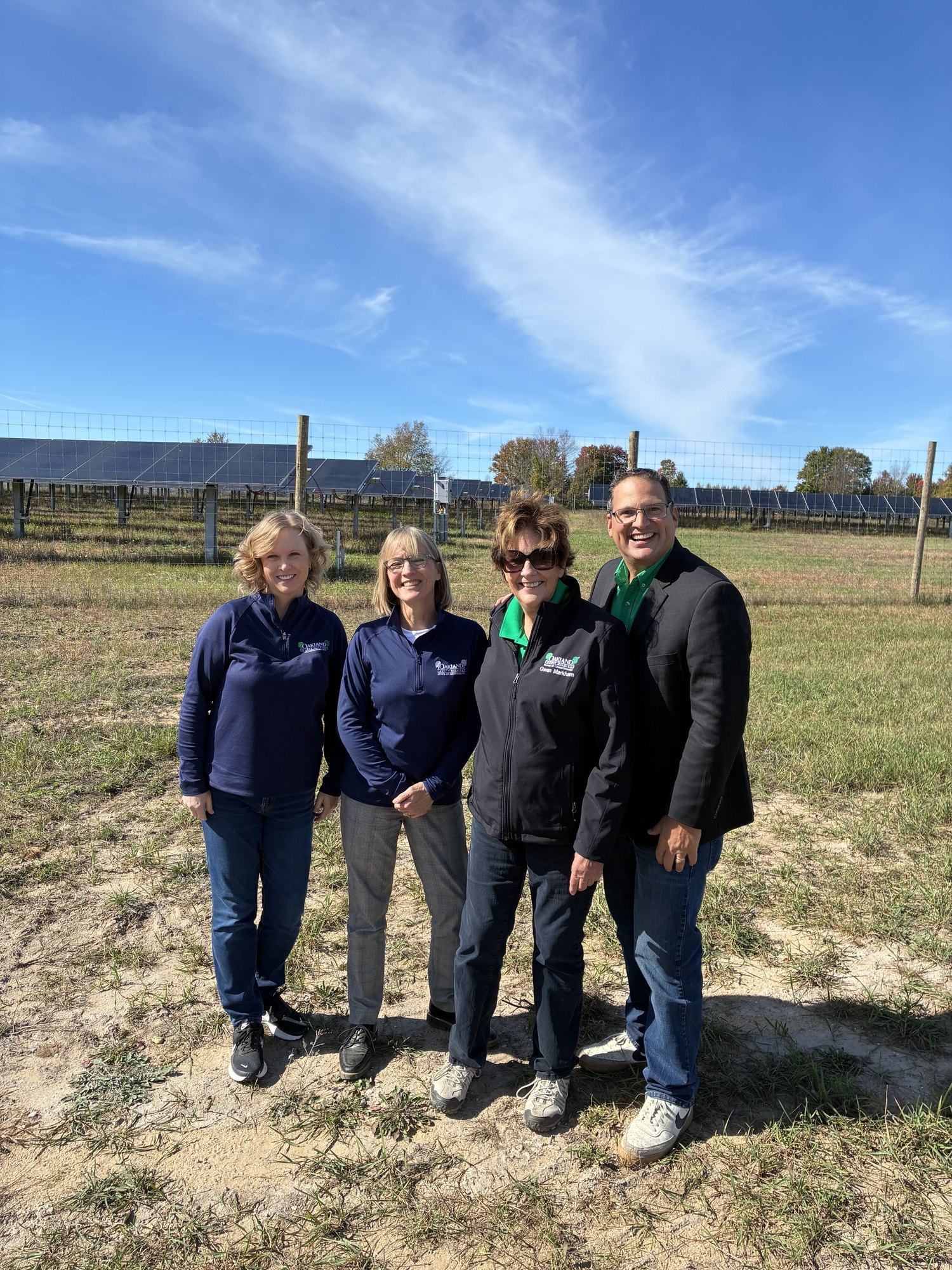 Four people stand outdoors in a field with solar panels visible behind a fence in the background.