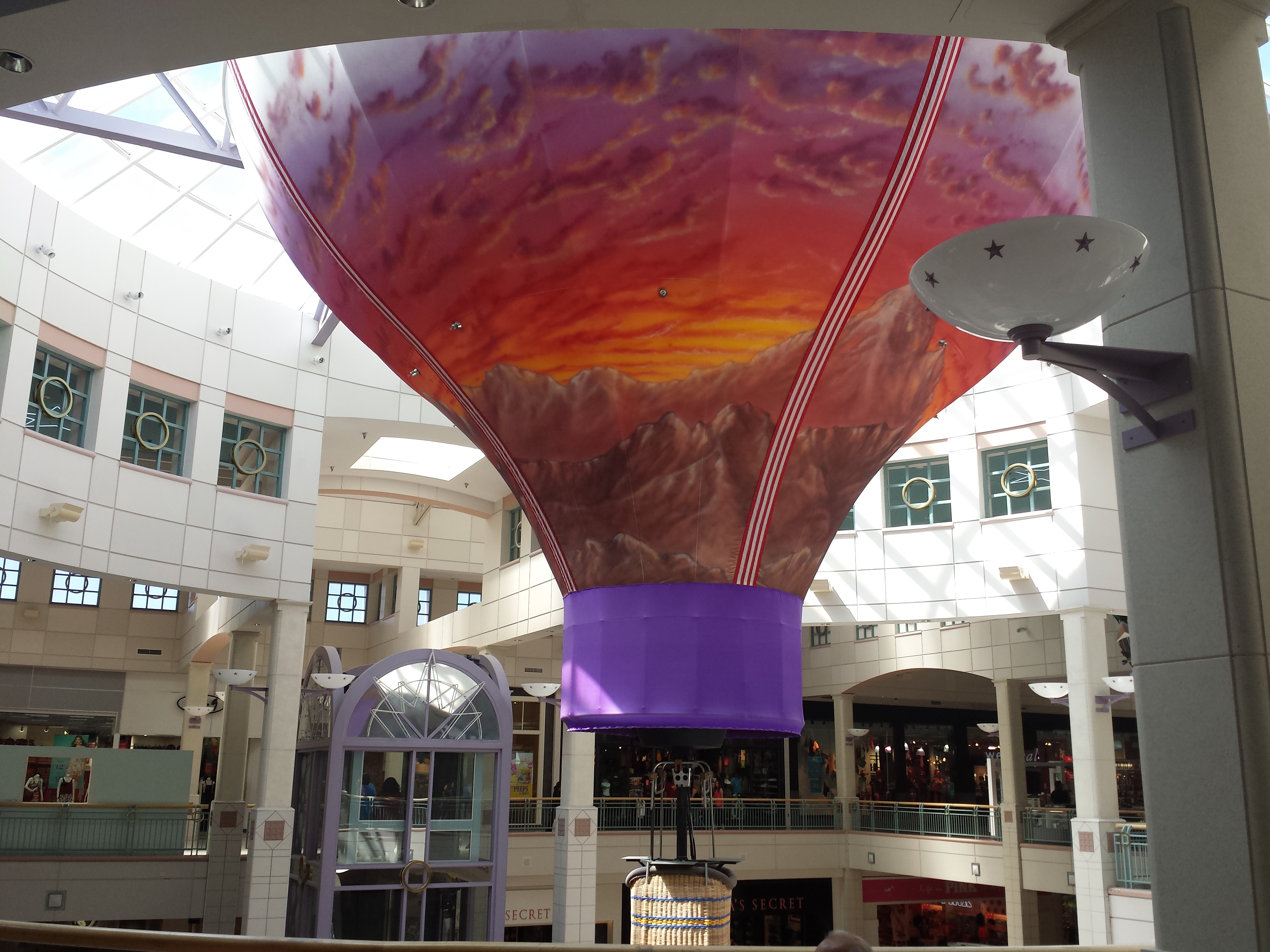 Multi-level mall atrium with a large colorful hot air balloon decoration suspended from the ceiling.