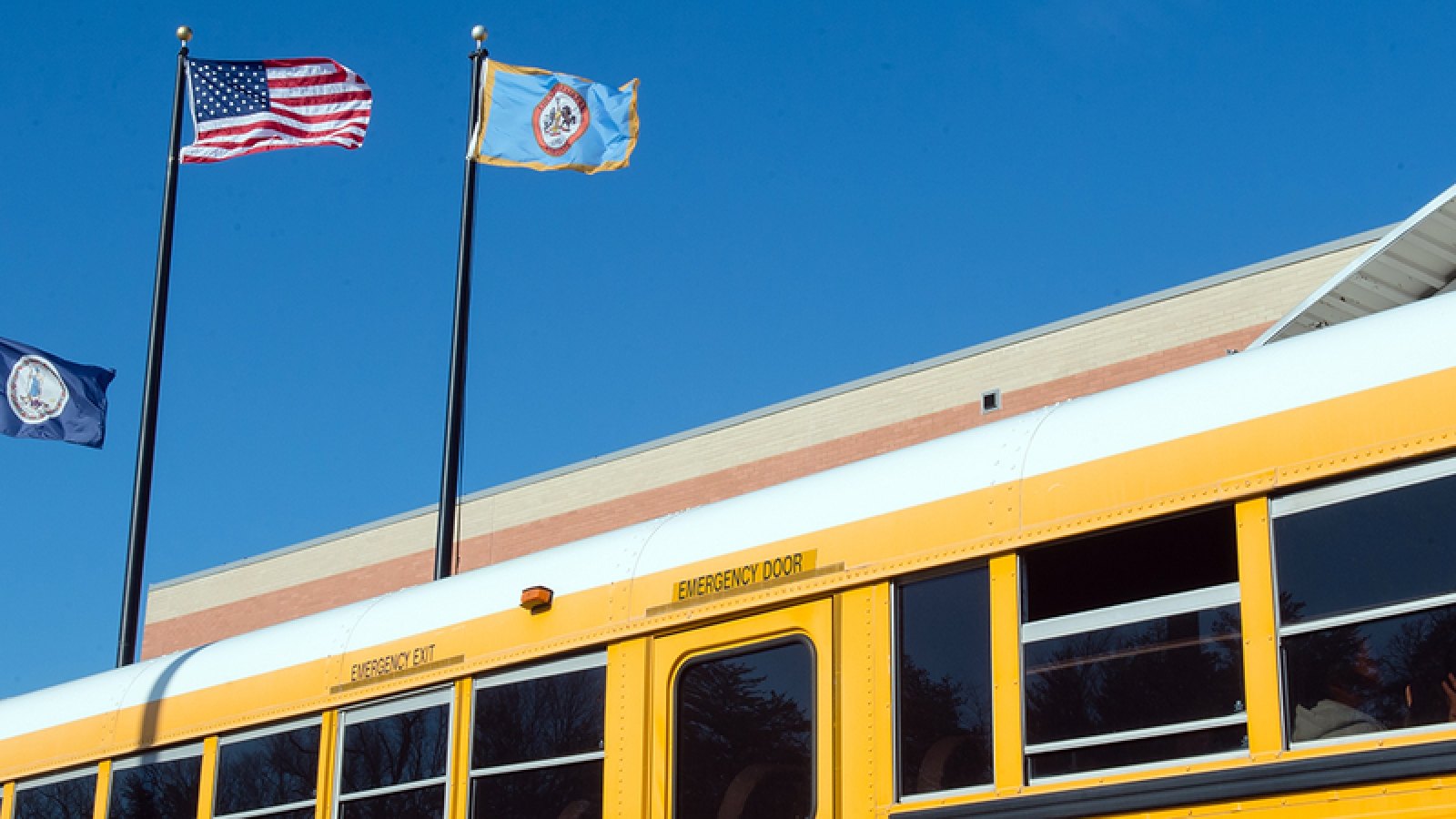 Yellow school bus parked in front of a school building with three flags flying.