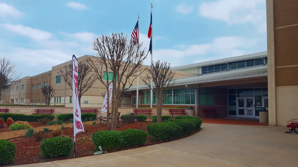 Front entrance of a large tan brick high school with two flagpoles.