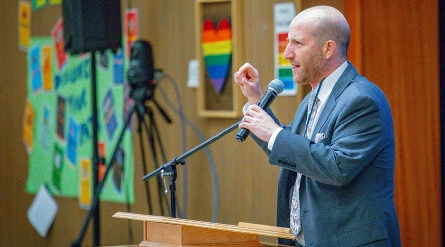 A man in a blue suit speaks at a podium microphone, gesturing with a raised fist, with colorful student artwork on the wall behind him.